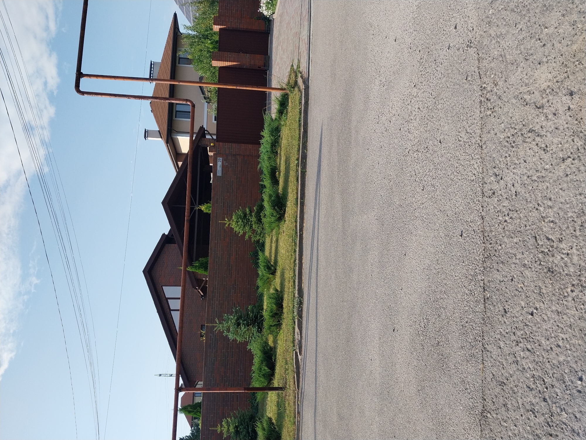 The image depicts a street view in the city of Doslidne, Ukraine. In the foreground, there is a paved road. Behind the road, a low-lying green lawn is visible, interspersed with several small evergreen trees and manicured bushes. A brick wall borders the property behind the lawn. The wall is topped with a continuous brown metal railing. Beyond the wall, two houses are partially visible. The house on the left has a dark brown brick facade and a steeply pitched dark roof with triangular windows. The house on the right is lighter in color with a brown roof and visible chimneys. A brown metal gate is present in the wall, and part of a paved driveway leads to it. The sky above is blue with some scattered clouds. Overhead, power lines are visible against the sky. A metal pole with a large pipe structure is prominent in the mid-ground, extending upwards from the foreground into the frame.