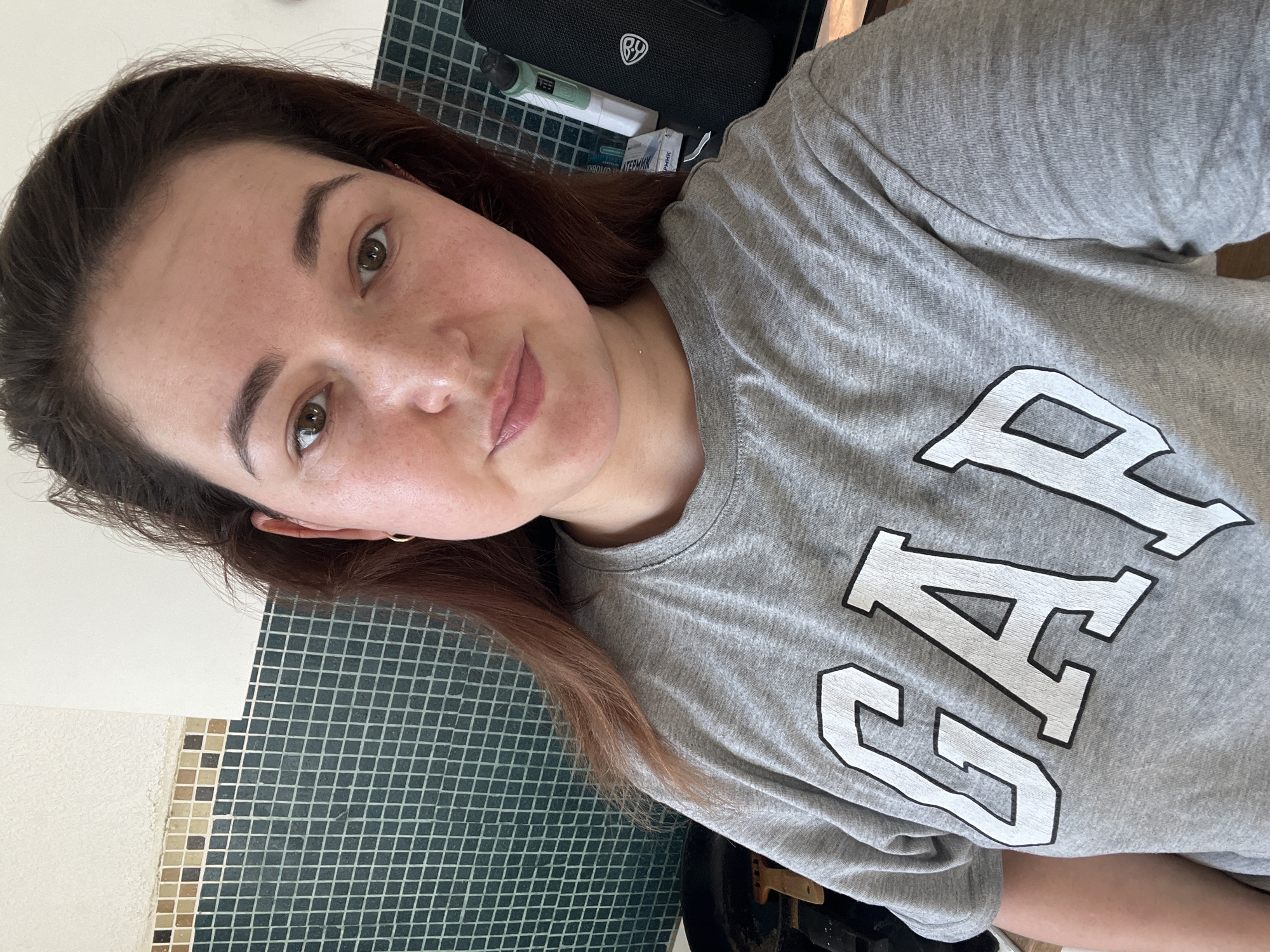 The media file is a selfie of a young woman. She is wearing a grey t-shirt with the word "GAP" printed on it in large white letters outlined in black. Her hair is dark brown and falls past her shoulders. She has light skin with freckles and is looking towards the camera with a neutral expression. She is wearing a small gold hoop earring.
The background shows a tiled wall, a black object with a logo and some electronic devices, and a grid-like surface. The lighting suggests it is daytime.
The visible text on the t-shirt says "GAP". There is also some text on a small white packet in the background, which appears to be in Cyrillic script, potentially related to medicine or cosmetics.
The image is taken indoors, likely in a bathroom or vanity area, given the tiled wall and the presence of personal items. The overall mood is casual and relaxed. The context is Chelyabinsk, Russia.