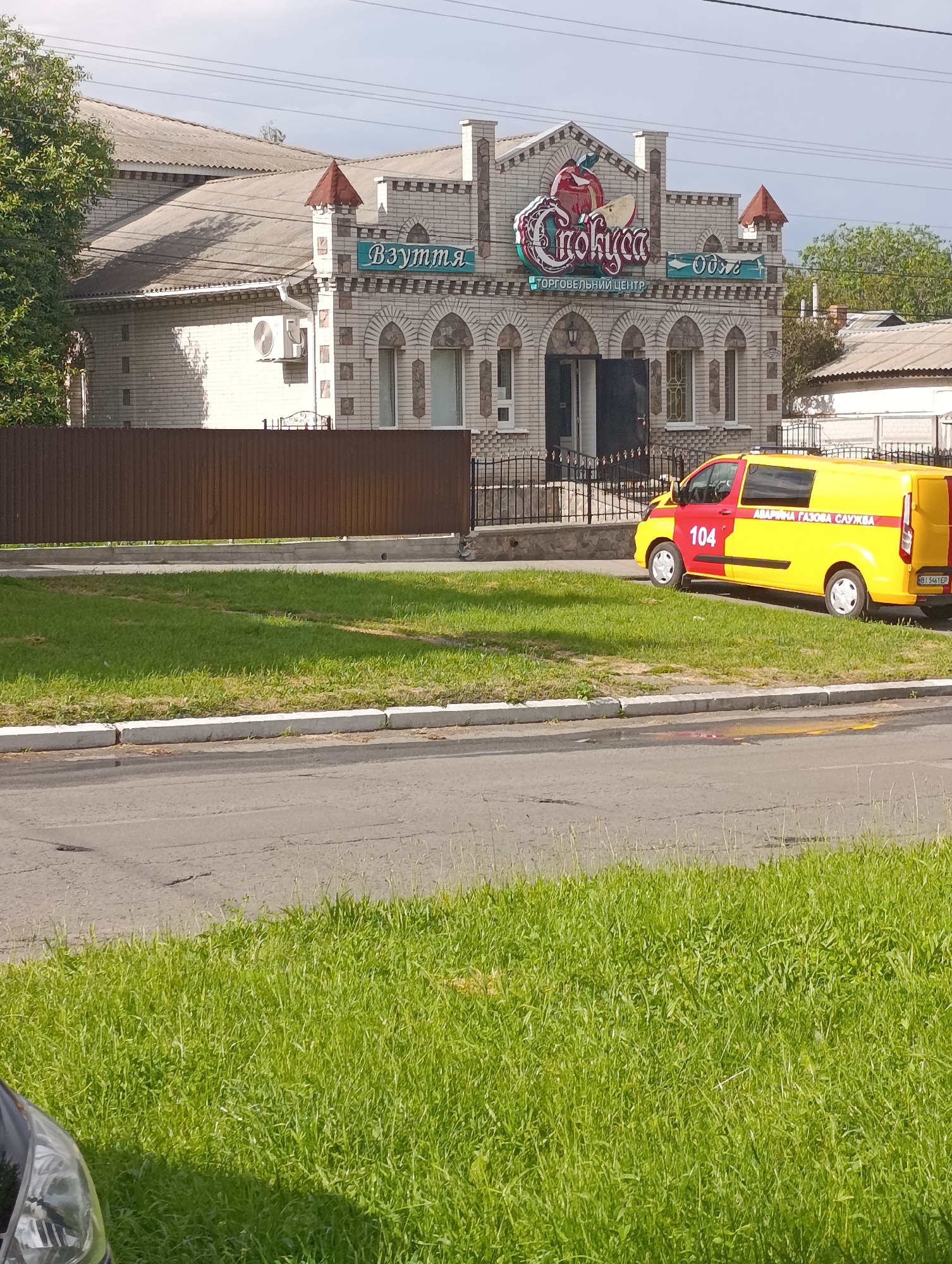 The image shows a street scene in the city of Khorol, Ukraine. In the background, a two-story building with a white brick facade and a grey corrugated roof is visible. The building has arched windows and decorative brickwork. Above the entrance, a neon sign displays the text "Слобода" with an apple motif, and to its right, another sign reads "Одеж". Below the "Слобода" sign, text reads "ТОРГОВЕЛЬНИЙ ЦЕНТР". To the left of the building, a green tree provides shade. In the mid-ground, a red and yellow service van with the number "104" on its side is parked on the grass next to the road. The van also has text that appears to read "АВАРІЙНА ГАЗОВА СЛУЖБА". A brown corrugated metal fence runs in front of the building. The foreground consists of a paved road and a strip of green grass.