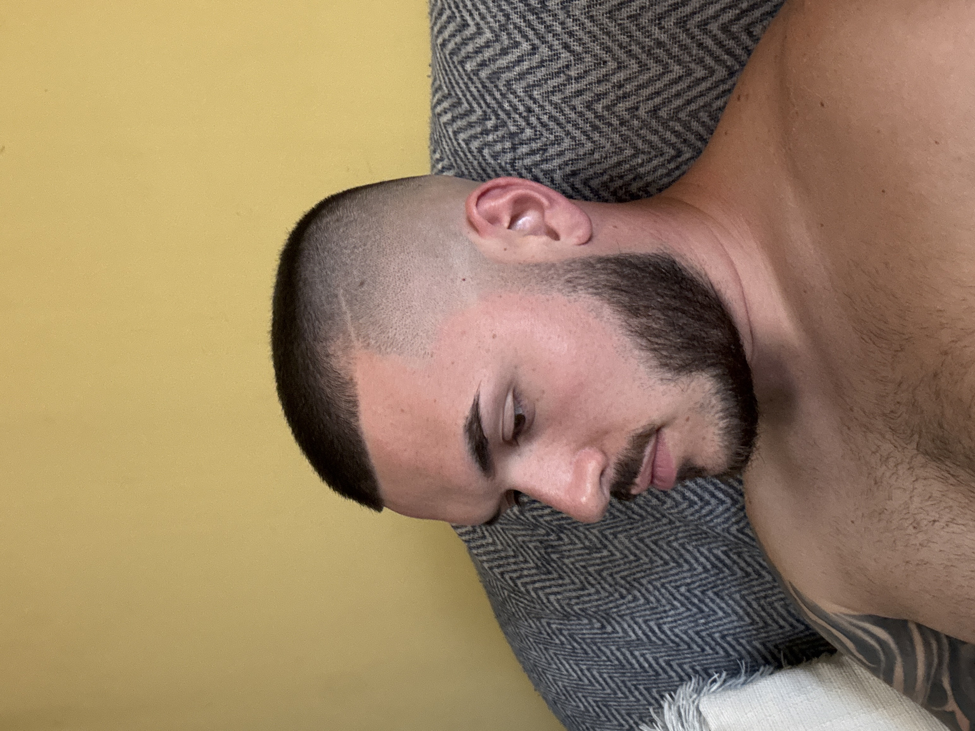 A close-up shot shows a light-skinned male with a short, dark haircut and a beard, resting on a gray and white patterned textile. The man's eyes are closed, and he appears to be sleeping or resting. Part of his bare chest and shoulder is visible. The background consists of a solid light yellow wall. There is no indication of the city of Pazardzhik, Bulgaria in the image.