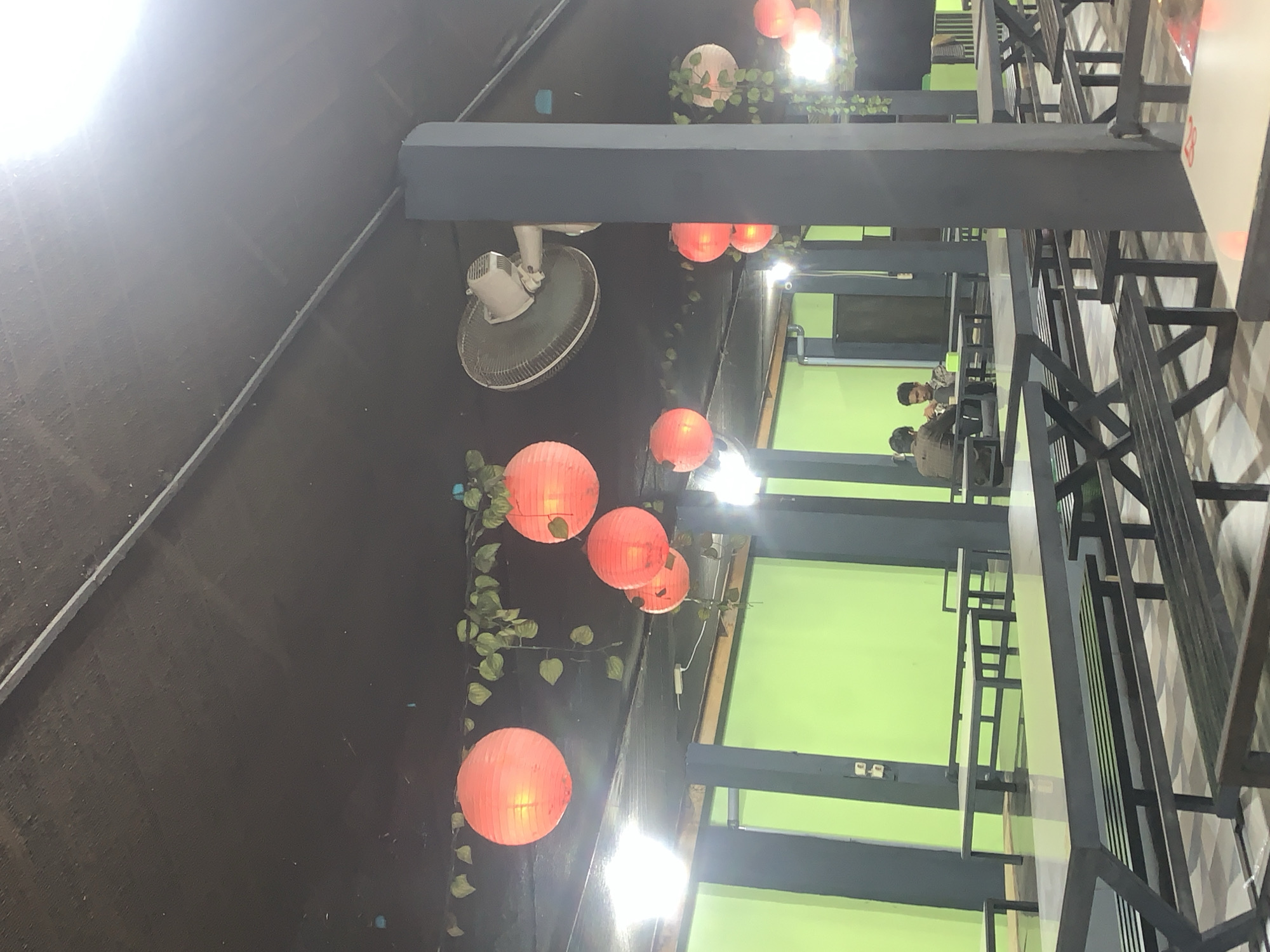 The image depicts an outdoor seating area at a restaurant in Palohlada, Indonesia. The scene is illuminated by several bright orange paper lanterns hanging from the ceiling, interspersed with artificial green vines. A large oscillating fan is mounted on the dark ceiling, and industrial-style lighting fixtures are visible.
The seating area consists of numerous long, grey tables with matching benches, arranged in rows. The tables have a reflective surface, suggesting they are made of a material like polished stone or laminate. The benches are constructed from dark wood slats.
In the background, two young men are seated at a table, facing each other. One man is wearing a dark jacket, and the other is wearing a lighter colored long-sleeved shirt. They appear to be engaged in conversation or a meal. The walls behind them are painted a vibrant lime green. A dark grey pillar is prominently positioned in the foreground, partially obscuring the view of the seating area. The overall atmosphere suggests a casual dining environment.