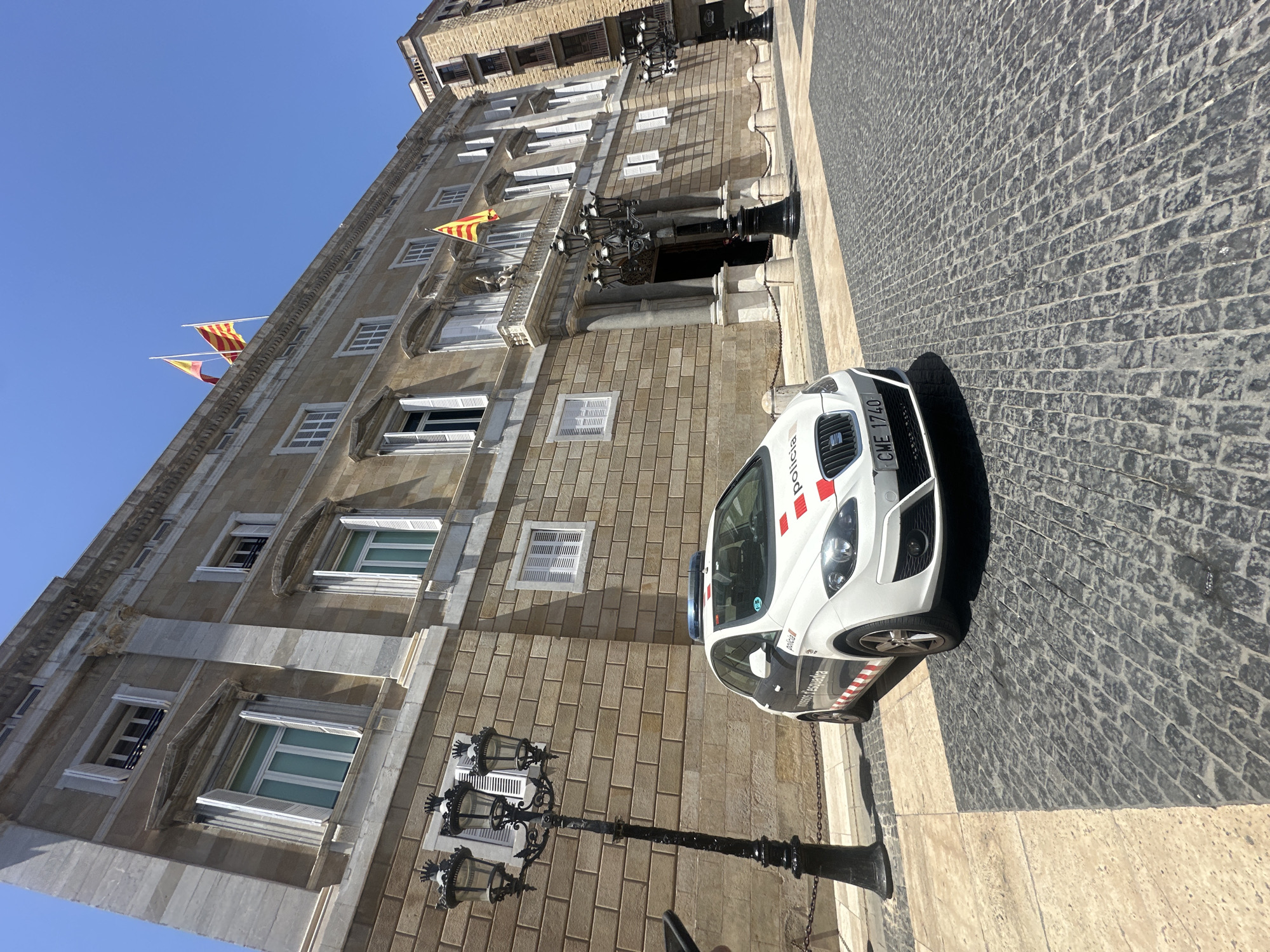 The image shows a white police car parked on a cobblestone street in front of a large, ornate building. The building is made of stone and has multiple windows with white shutters. There are flags flying from the top of the building, one of which is a Catalan flag. The police car has "policia" written on its side and appears to be a Seat Leon. The street is paved with dark gray cobblestones, and there are two black lampposts with multiple lights on the left side of the image. The sky is clear and blue. The scene appears to be located in a public square or plaza in Barcelona, Spain.