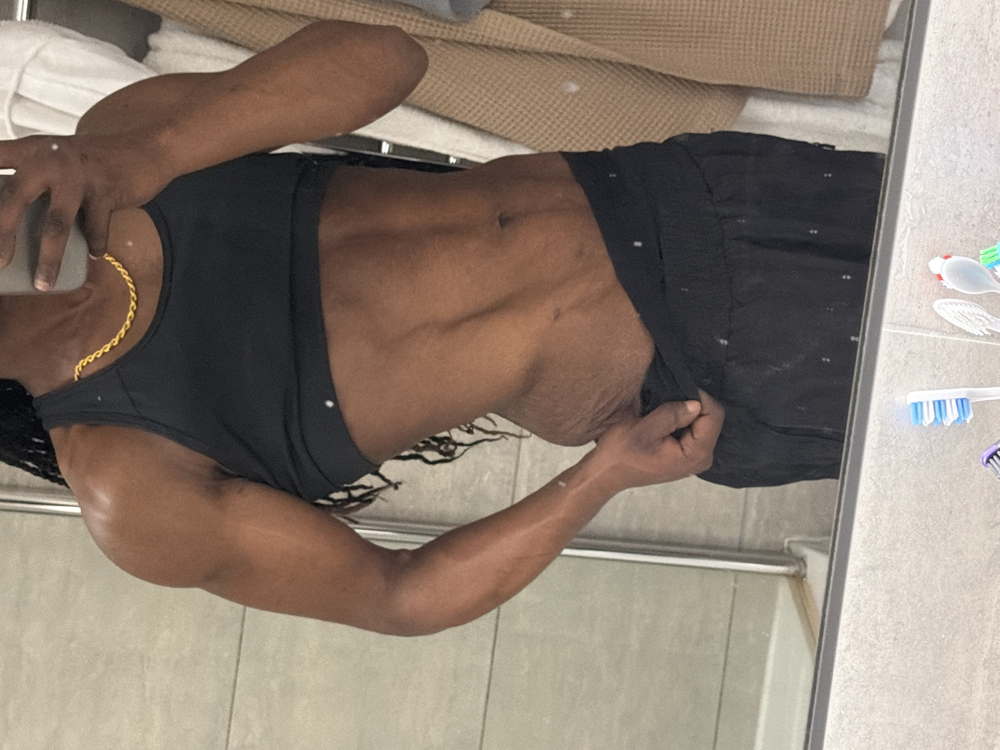 The image displays a person taking a mirror selfie in an indoor setting, likely a bathroom, with a provided context of London, United Kingdom.
The primary subject is a person with dark skin, positioned sideways to the camera's perspective. They are wearing a black sports bra or crop top and dark-colored shorts. A gold chain is visible around their neck. Their left hand, holding a phone, is raised towards their head, while their right hand is touching or adjusting the hem of their shorts. The person exhibits a muscular physique, with visible abdominal definition and toned arms. Their hair appears to be in braids or dreadlocks, partially visible on the left.
The background of the mirror reflection shows a light-colored, possibly waffle-textured mat or rug and a white towel or fabric. The floor in the reflection and on the right side of the mirror consists of light grey or beige tiles. A metal bar or railing is visible horizontally in the lower section of the mirror.
On a surface to the right of the mirror, likely a bathroom counter, several toothbrushes are present. One features a blue and white head, and another has a purple head. A small, clear object, possibly a holder or suction cup, is also visible near the toothbrushes.
The scene suggests a personal moment of self-documentation. The time of day is not discernible, as the lighting appears to be artificial. There is no visible text in the image.