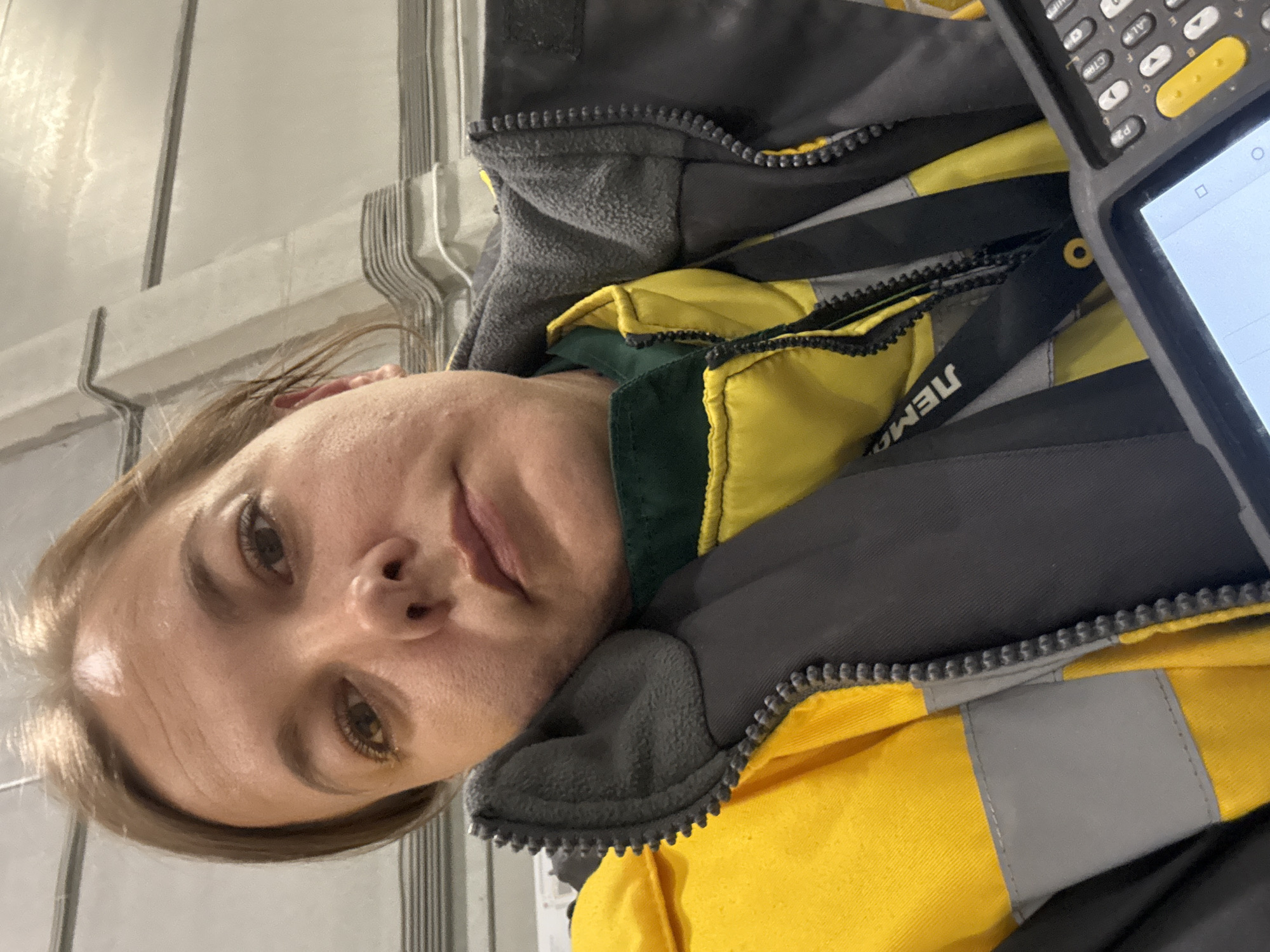The image, originally rotated 90 degrees clockwise, depicts a person, likely a woman, in an indoor industrial setting. She has light brown hair, fair skin, and brown eyes, looking directly at the camera with a neutral to slightly contemplative expression.
She is wearing a work uniform consisting of a gray and yellow high-visibility jacket with a dark green underlayer visible at the collar. The jacket features prominent zippers and hints of reflective stripes. On a black strap or lanyard visible across her chest, the text "JEMO" (or similar, partially visible) is printed.
In the upper right corner of the image, a handheld electronic device, resembling a scanner or mobile computer, is visible. It features a physical keypad with various buttons, including 'SHIFT', 'ALT', 'ESC', 'CTWL', and alphanumeric keys 'A' through 'F', along with yellow accent buttons. The device's screen displays a digital interface, possibly showing a search bar or an open application.
The background consists of light-colored, possibly metallic or concrete, walls or structures with vertical and horizontal lines, suggesting a warehouse, factory, or similar operational environment. The presence of the work attire and the scanning device indicates the person is engaged in a work-related activity, such as inventory management or logistics. The location context is Lupanovo, Russia.