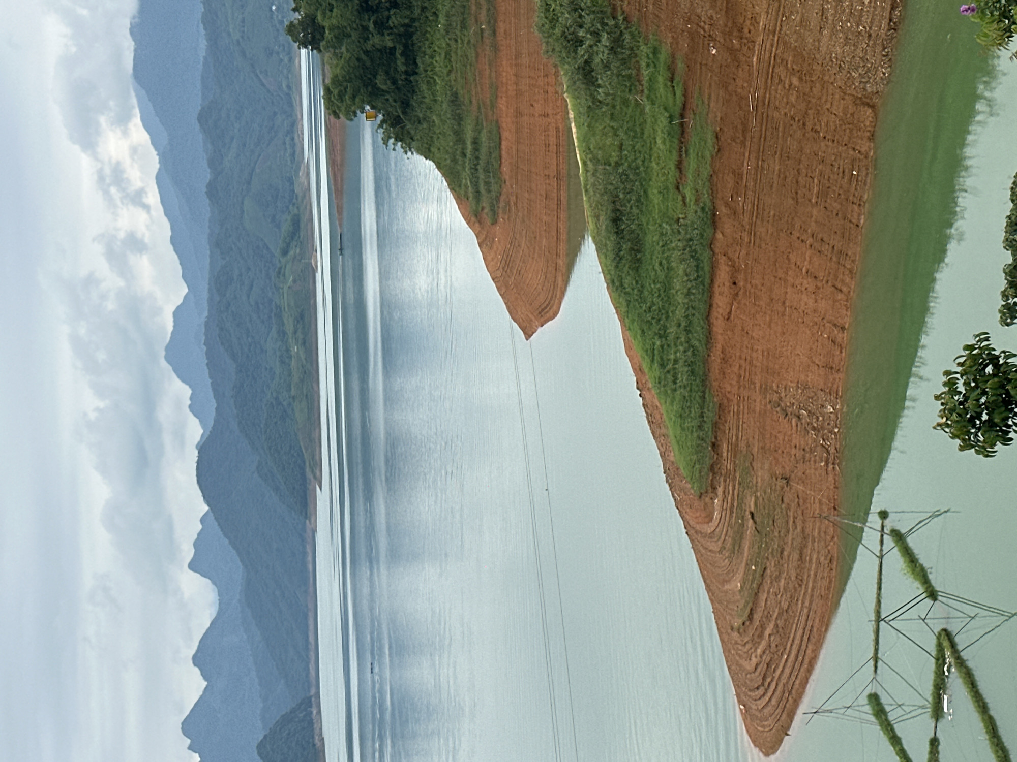 The image depicts a wide, calm body of water, likely a reservoir or lake, with a distinct shoreline characterized by reddish-brown soil layered in terraces. Lush green vegetation covers some of these terraces and the hills rising beyond. In the background, a series of misty, blue-toned mountains form a dramatic horizon under a cloudy sky. The water itself is a pale blue-green, reflecting the overcast conditions. In the foreground, partially submerged structures with green vegetation are visible, forming geometric patterns within the water, suggesting some form of aquaculture or water management system. A few faint power lines stretch across the water in the mid-ground. The scene appears to be a natural landscape with some human intervention, possibly in or near Bản Phúc, Vietnam, given the mountainous terrain and water body.