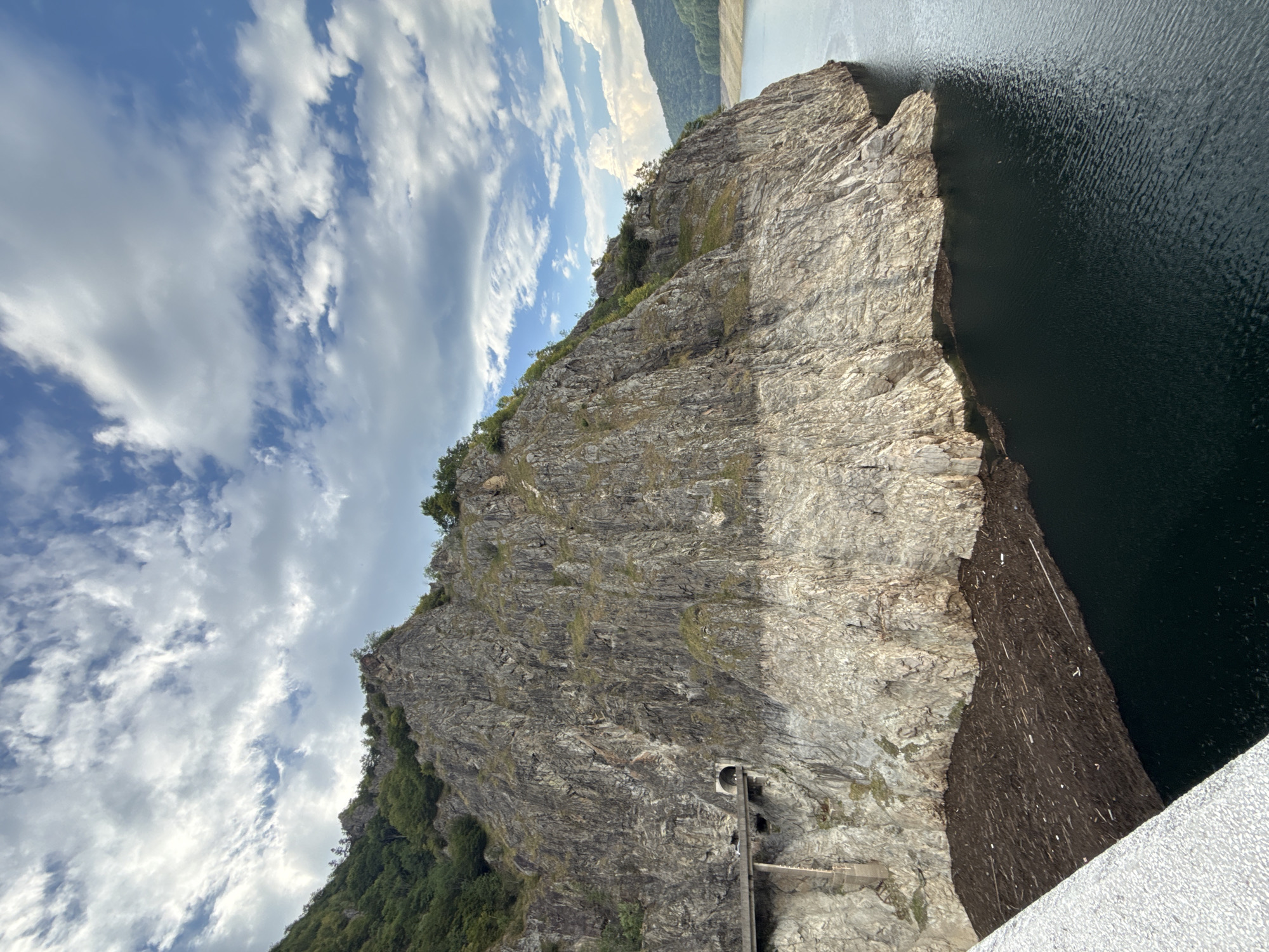 This image captures a scenic view of the Vidraru Dam in Arefu, Romania. The dam, a significant concrete arch dam on the Argeș River, is situated in a mountainous region characterized by steep, rocky cliffs. The scene showcases the impressive scale of the dam and the surrounding natural landscape.
The upper portion of the image is dominated by a vast sky filled with fluffy white clouds against a backdrop of blue. The clouds are scattered and appear to be moving, suggesting a dynamic weather pattern.
Below the sky, the rugged face of the mountain that the dam is built into takes center stage. The rock formations are varied, with different textures and colors indicating age and geological composition. The upper parts of the cliffs are covered in lush green vegetation, with trees and shrubs clinging to the slopes, adding a vibrant contrast to the grey rock.
A notable feature carved into the side of the mountain is a tunnel, presumably part of a road or access route. A concrete structure, likely a bridge or support for the road, is visible leading to the tunnel entrance, and several vehicles can be seen on the road.
The lower part of the image reveals the dark, still waters of the reservoir behind the dam. A significant amount of debris, primarily composed of logs and branches, is accumulated along the edge of the water where it meets the rocky shore. This accumulation suggests a recent event, possibly related to the dam's operation or a natural occurrence like a flood or storm. The water's surface is calm, reflecting the surrounding landscape and sky.
In the distance, rolling hills covered in dense green forests can be seen, extending towards the horizon. These hills contribute to the overall sense of a remote and natural environment. The foreground shows a portion of what appears to be the top of the dam structure itself, a textured, light-colored surface which contrasts with the natural elements.