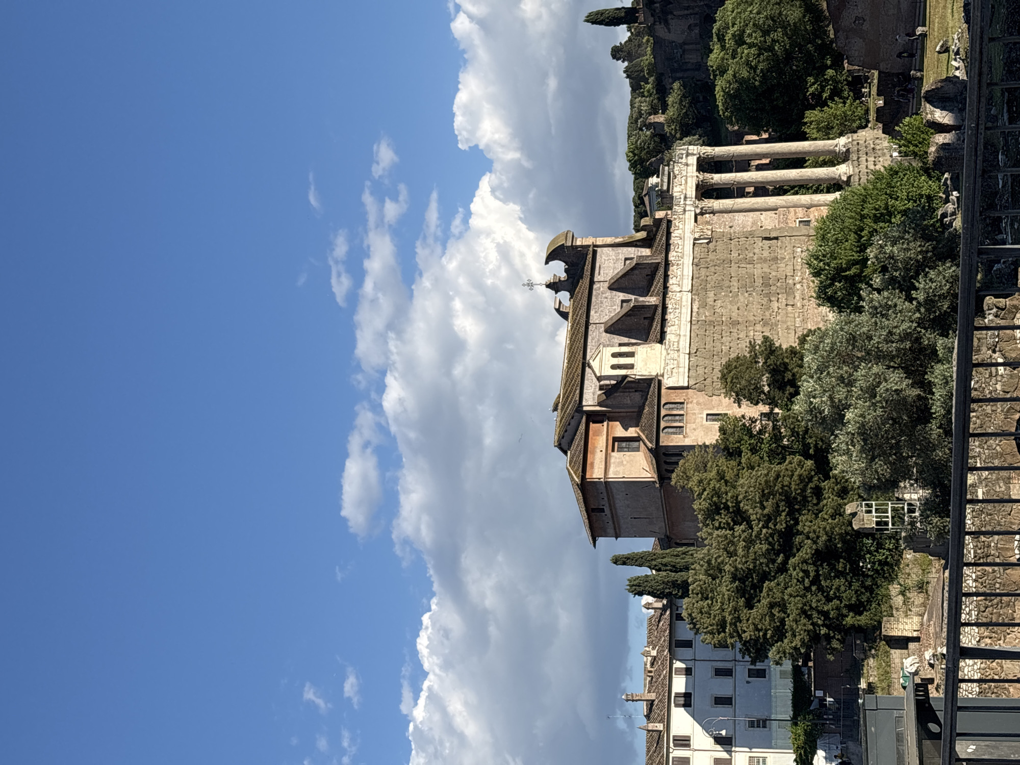 The image depicts a view in Rome, Italy, featuring the Basilica di Santa Francesca Romana. The church is constructed with stone walls and a tiled roof, topped with a cross. Adjacent to the Basilica are partial views of the Roman Forum ruins, characterized by stone columns. The surrounding environment is lush, with green trees and plants. In the foreground, there is a metal fence or railing. The sky is visible with a mix of blue sky and white clouds. The buildings across the way are white with square windows. The basilica has an orange stone section.
