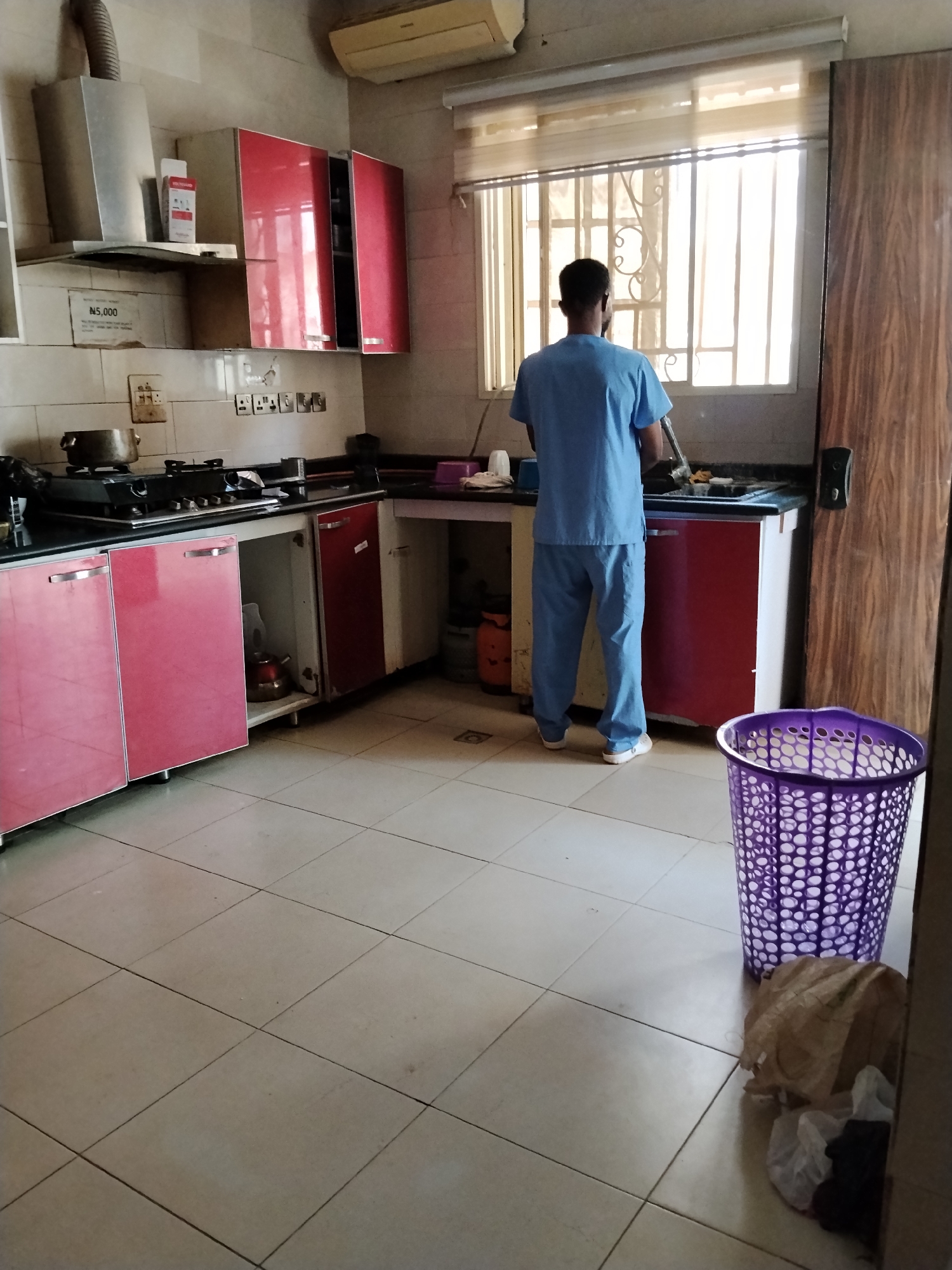 In a brightly lit kitchen, possibly in Gwarinpa, Nigeria, a person wearing blue scrubs stands at a sink area, facing away from the camera. The person appears to be engaged in washing or preparing something. The kitchen features red cabinets above and pink cabinets below the countertop. A stainless steel range hood is visible above the stove, which has a pot on it. The floor is tiled with square beige tiles. A large purple laundry basket sits on the floor near the right side of the frame, beside a crumpled brown paper bag and some white plastic bags. The room is well-lit by natural light streaming through a window covered with blinds.