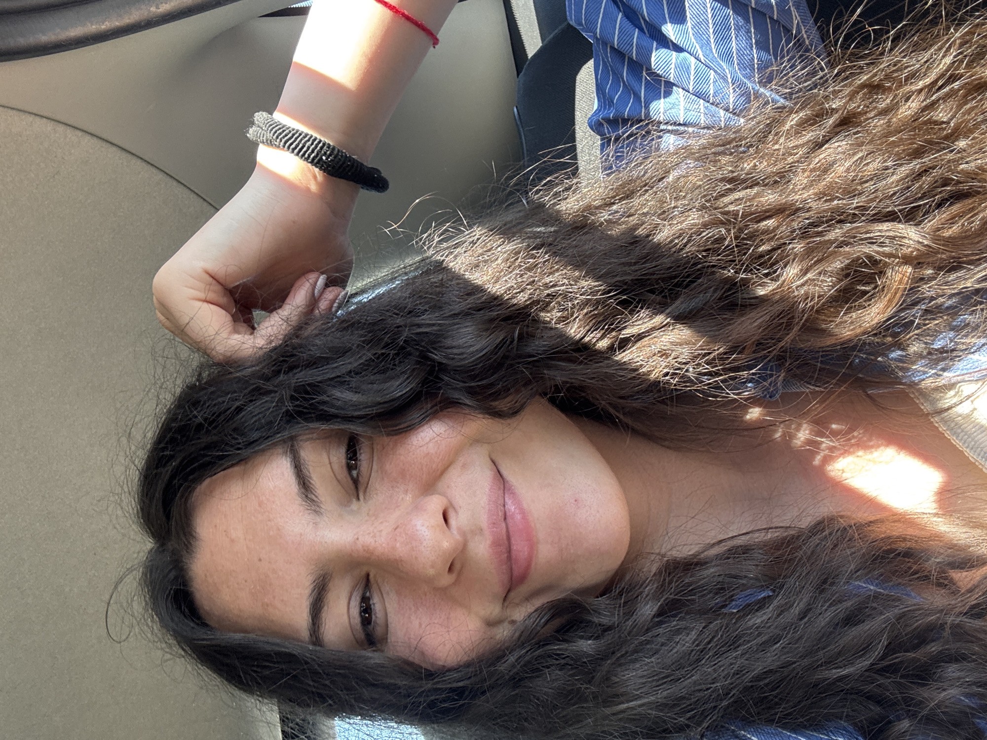 The image is a selfie of a woman with dark hair, lying down, possibly in a car. She is wearing a blue and white striped shirt, and has two bracelets on her left wrist, one red and one black. The location appears to be Unknown, Unknown. The lighting indicates it is likely daytime.