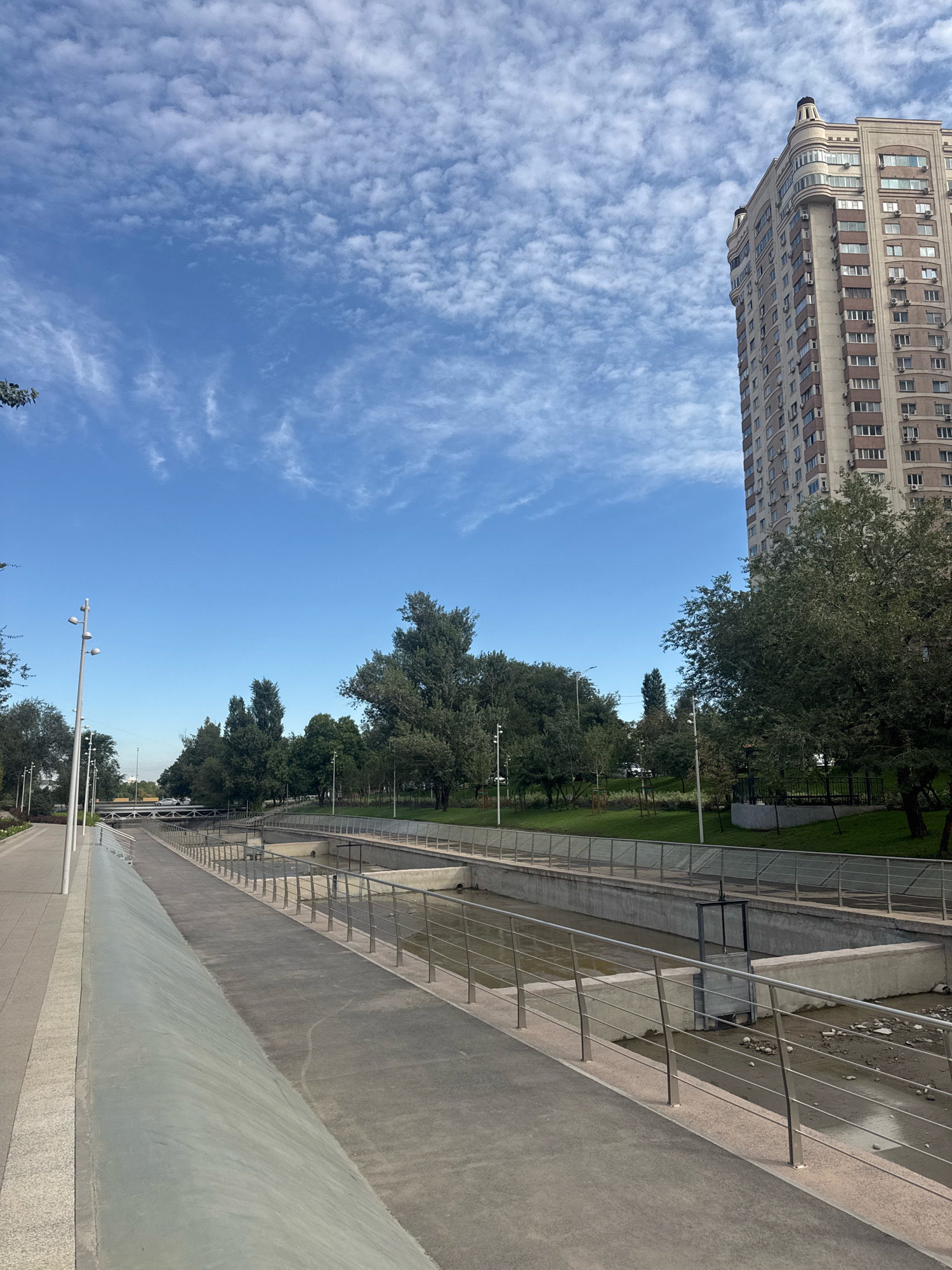 The image captures a daytime view of the embankment of the Malaya Almatinka River in Almaty, Kazakhstan, near the intersection of Furmanov Street and Satpayev Avenue. In the foreground, a paved walkway runs alongside a concrete embankment, separated by a metal railing. The riverbed is visible, with shallow, murky water and scattered stones. Lush green trees and grass line the opposite bank, where a road with moving vehicles can be seen in the distance, as well as a pedestrian bridge. Dominating the right side of the frame is a tall, multi-story apartment building with a distinctive turreted roof, characteristic of the Stalinist Empire style architecture prevalent in the area. The sky is a clear blue, punctuated by scattered white, wispy clouds. The scene depicts an urban park area integrated with the river and a residential building, suggesting a developed urban landscape.