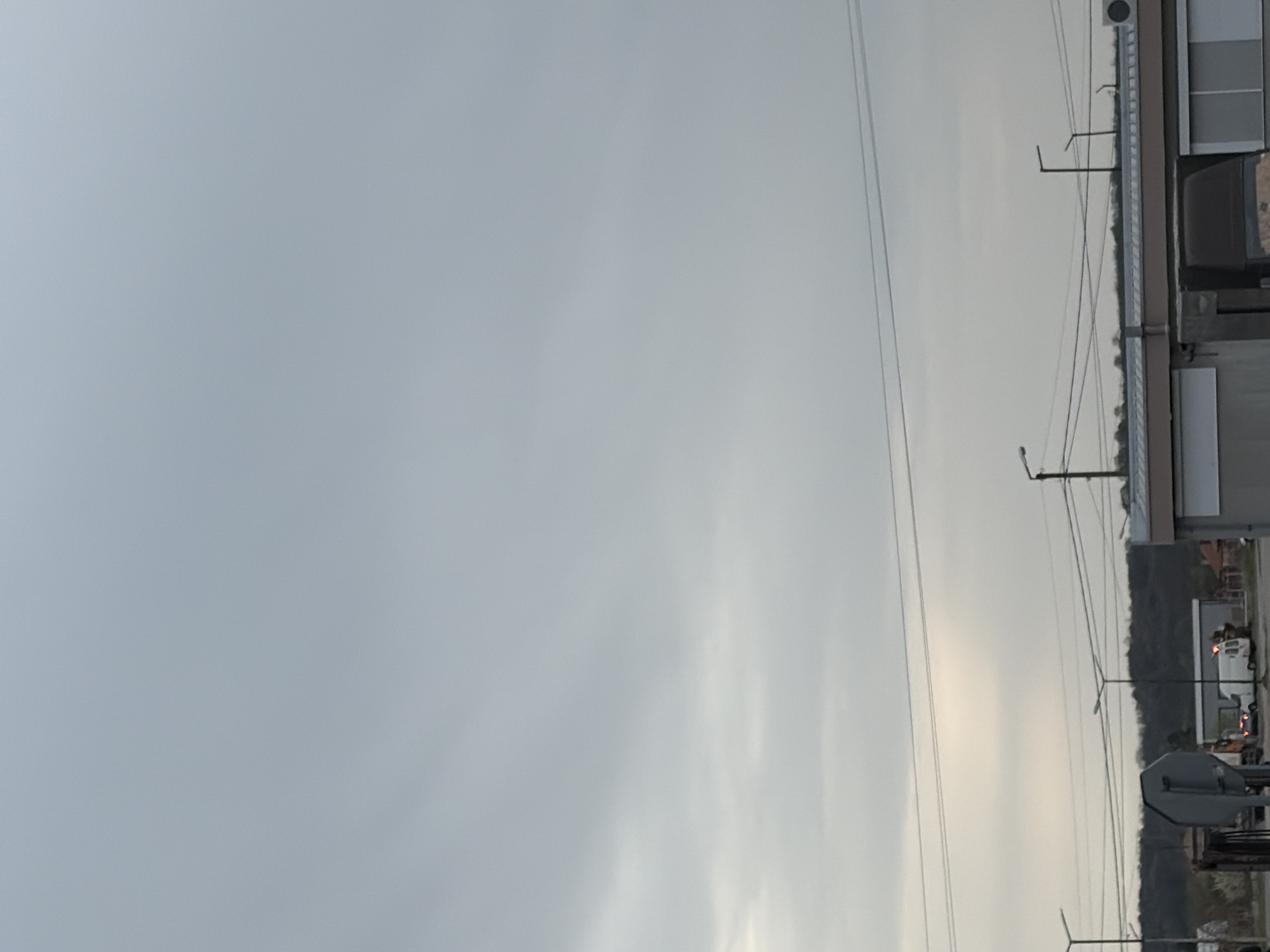 The image depicts a sky with overcast, grey clouds. The lower right side of the frame features power lines, utility poles, and parts of buildings. It's a view in Plodovitovo, Bulgaria. A treeline and parts of vehicles are visible in the distance, partially obscured by the utility infrastructure.