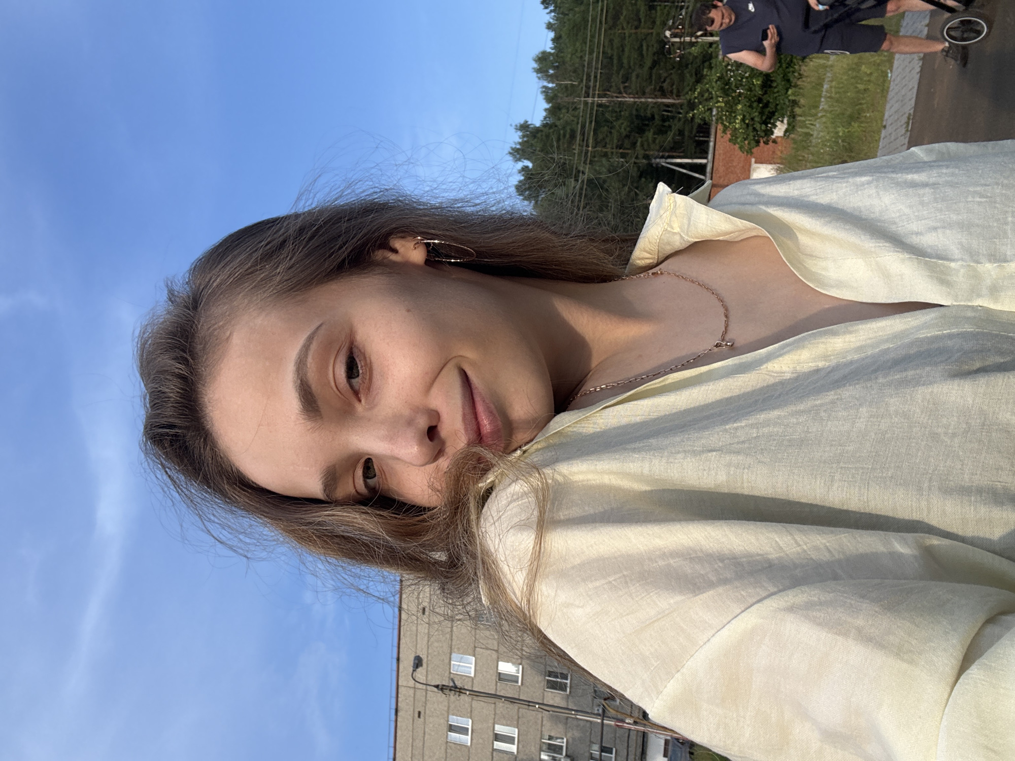 A young woman with long, light brown hair is taking a selfie outdoors. She is wearing a light yellow button-down shirt and a delicate gold necklace. She is smiling at the camera, and her hair is blowing in the wind. In the background, a grey apartment building with white-trimmed windows stands on the left, and a line of trees is visible on the right. A man is standing in the background on the right, looking at his phone. The scene appears to be in a residential area in Noviy, Russia, possibly near a park or a communal outdoor space. The sky is a clear blue with a few wispy clouds. The lighting suggests it is late afternoon or early evening.