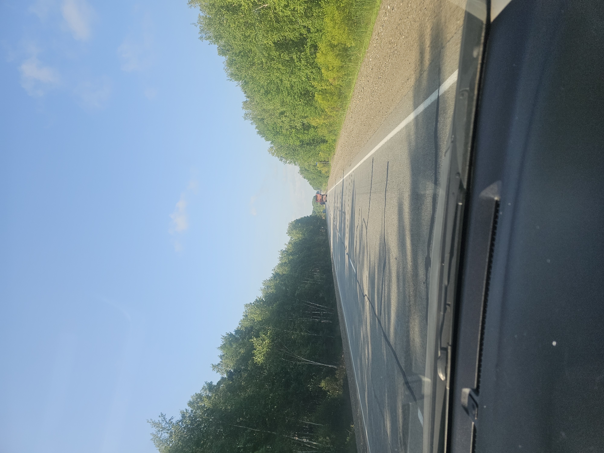 The image shows a view from inside a vehicle driving on a paved road lined with trees. The road has lane markings and appears to be a highway. In the distance, there are two trucks on the road. The sky is clear blue with a few wispy clouds. The environment appears to be a rural or suburban area, possibly in the city of Zoevka, Russia, given the provided context. The image is taken during daylight hours, and the lighting suggests it could be morning or late afternoon. There are no visible people in the image, and the focus is on the road and the surrounding landscape. The interior of the vehicle, including the dashboard and windshield wipers, is visible in the foreground, indicating the perspective is from the driver's or passenger's seat.