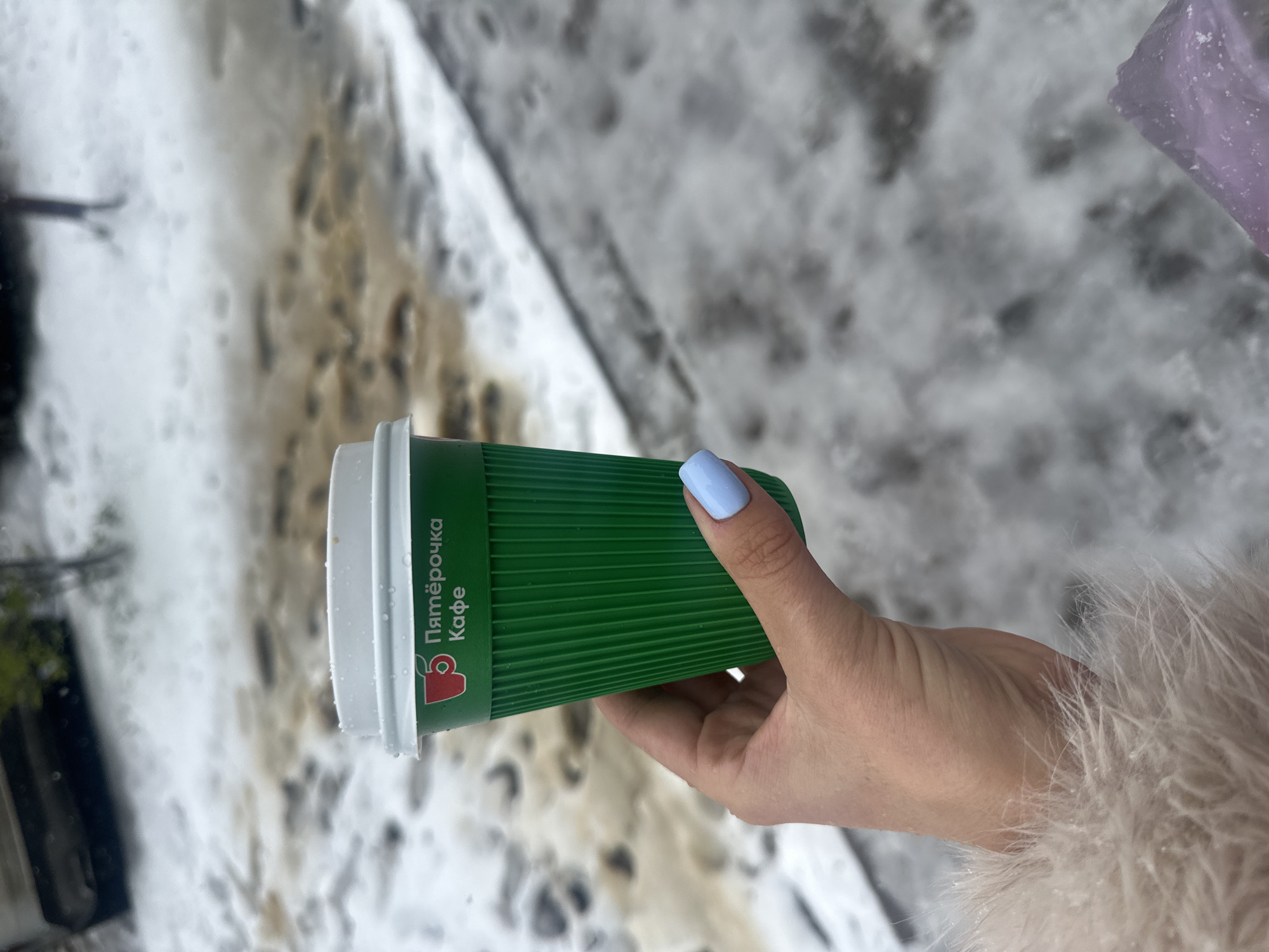A hand holds a green "Pyaterochka Cafe" coffee cup. The hand has pale skin and a fingernail painted light blue. The person is wearing a light fur-like jacket. The background shows a snowy, slushy area, likely a road or sidewalk. It is Krasnogorsk, Russia.