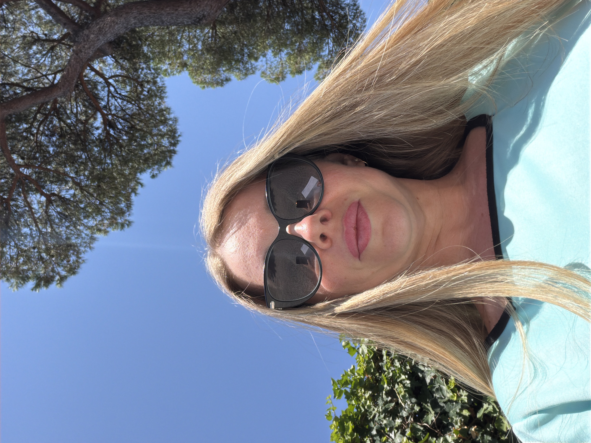 A low-angle, medium shot captures a woman with long blonde hair and sunglasses against a bright blue sky. She is wearing a teal top with a black neckline. Lush green foliage and a portion of a tree trunk with branches are visible in the background. The scene appears to be outdoors, possibly in Rome, Italy, given the Mediterranean vegetation. The sun is strong, casting shadows on her face and clothing.