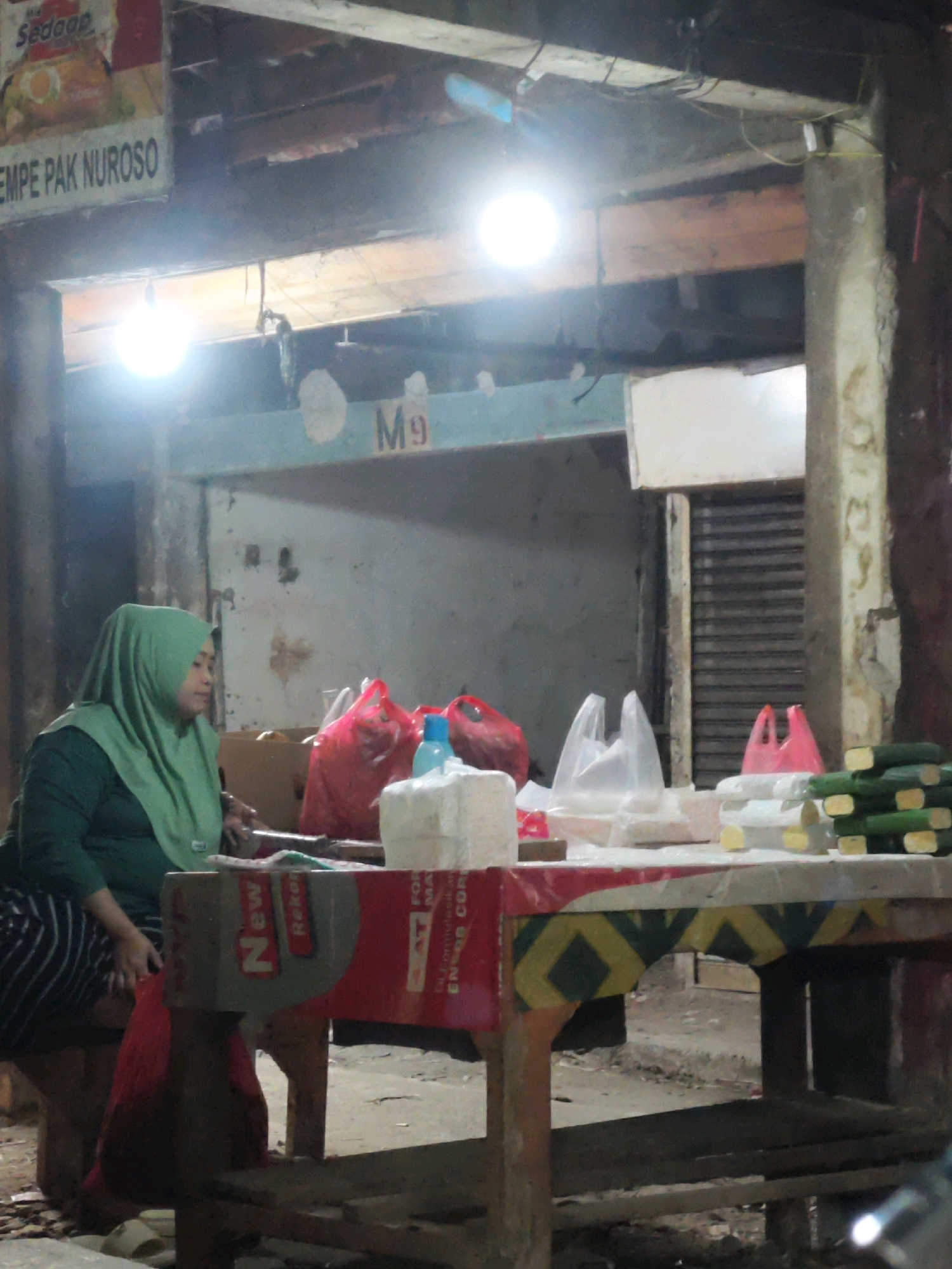 In a dimly lit outdoor market setting, likely in Karawang, Indonesia, a woman wearing a green hijab and a green top is seated at a table. The table is covered with a colorful, patterned cloth and displays various items for sale, including plastic bags filled with what appear to be goods, a white paper-wrapped item, and a stack of green vegetables that resemble cucumbers. Behind the woman, a sign reads "M9". The environment suggests a street-side stall with wooden structures overhead and visible electrical wiring, illuminated by bright, possibly artificial, lights. The overall scene depicts a vendor at her stall, possibly during evening hours given the lighting conditions.