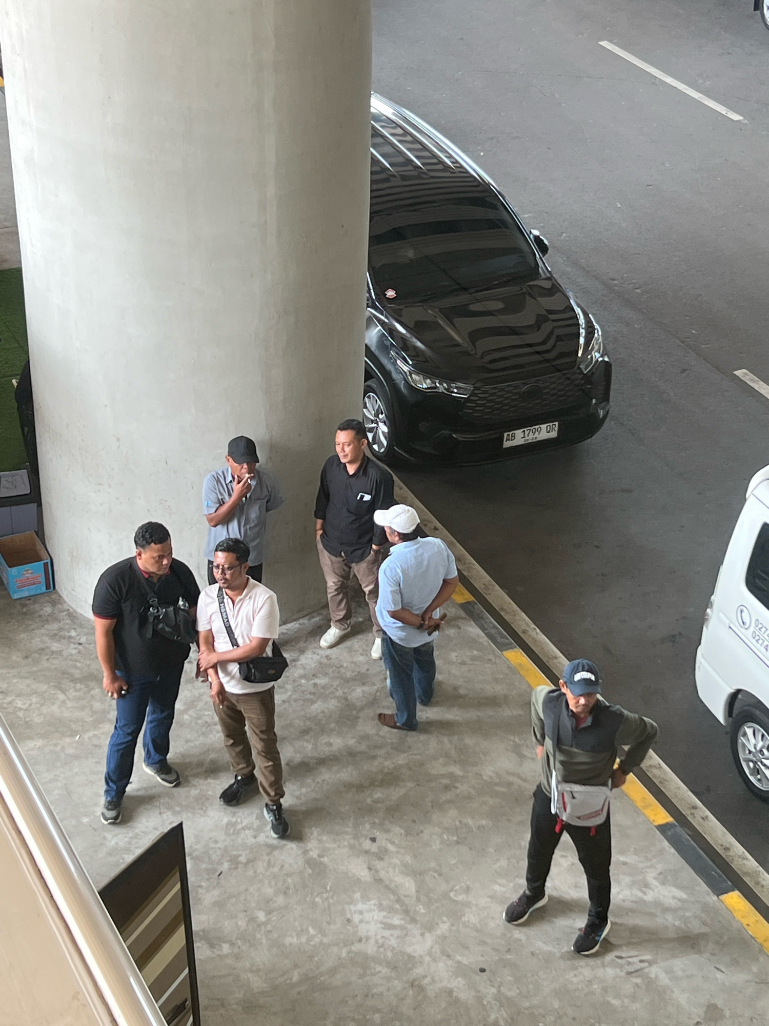 In Palihan, Indonesia, six men are standing near a large concrete pillar and a parking lot. One man in a grey shirt and black cap smokes a cigarette. Another man in a black shirt stands to his right. To their left, two men in black shirts and jeans converse. In the foreground, a man in a pink shirt and brown pants stands with a black bag, next to a man in blue jeans. A black car is parked behind them. A white van is parked further down the lot. The ground is concrete with yellow and black markings indicating a road.