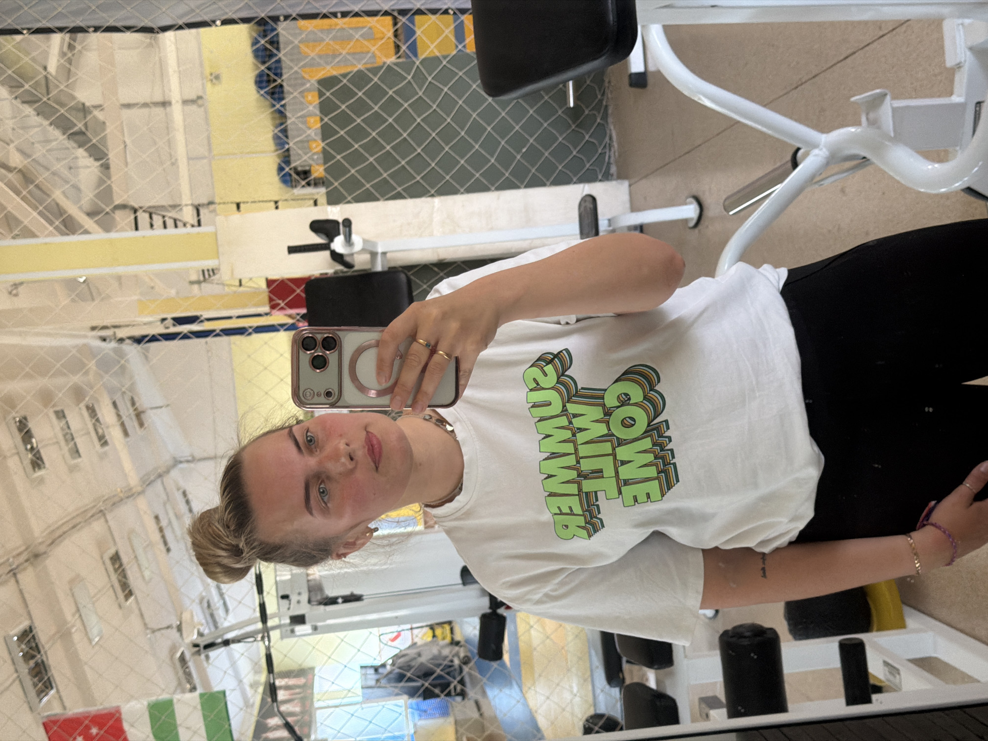 A young woman with blonde hair in a bun, wearing a white t-shirt with green text that reads "SUMMER WILL COME" and black pants, is taking a selfie in a gym. The gym appears to be indoors, with tiled flooring and metal equipment. In the background, there are reflections in a mirror showing more gym equipment, a flag with green and white stripes, and a blue and yellow net. The woman is holding a phone in her right hand and is wearing rings on her fingers. She has a subtle smile on her face. The scene is well-lit, suggesting it's daytime. The flag in the reflection may indicate a location outside of Russia, as the provided context indicates the city of Chaikovsky, Russia.