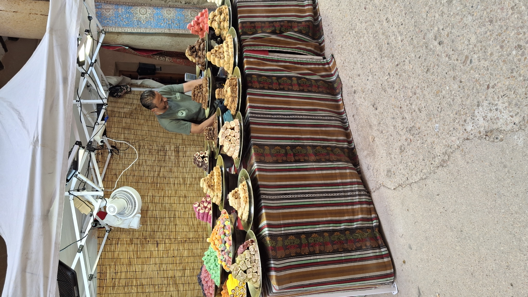 A man in a green t-shirt is standing behind a table laden with various types of Turkish delight. The table is covered with a striped cloth in brown, white, red, and green. The Turkish delight is arranged in piles on metal platters. The vendor appears to be restocking or arranging the sweets. Behind the table is a bamboo-like screen, and above it, a white tent structure with lighting. A small white fan is mounted on the screen. The scene appears to be an outdoor market stall. The image does not contain enough information to definitively determine the location in the city of La Fresneda, Spain.