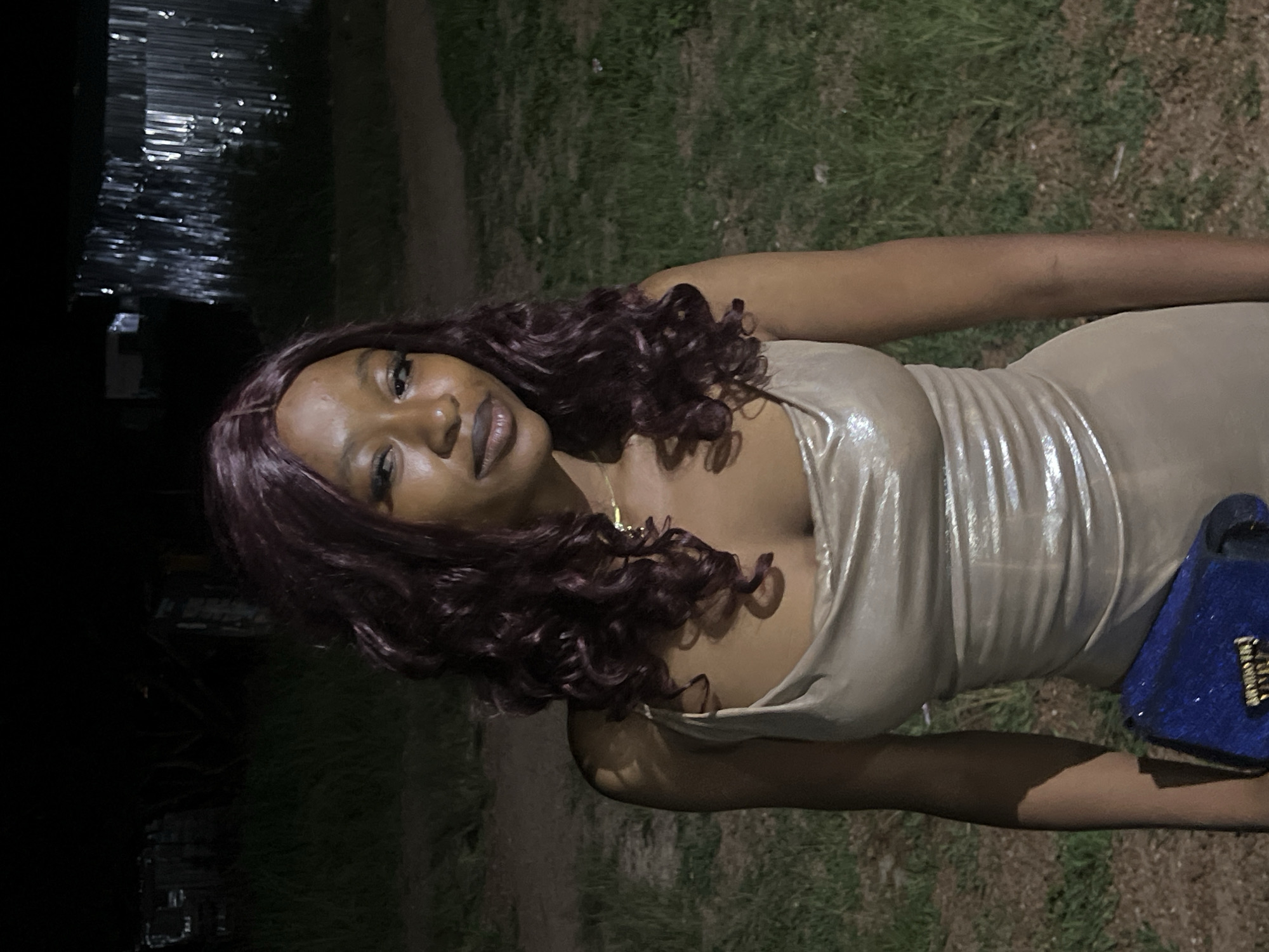 A young Black woman with curly, dark auburn hair and fair skin is pictured from the chest up, looking to her right with a slight smile. She is wearing a shimmery, champagne-colored slip dress with thin straps, and a delicate gold necklace. Her left arm is visible, holding a small, bright blue clutch bag. The background suggests an outdoor setting at night, with dimly lit grass and dry leaves on the ground. In the distance, there are structures, possibly buildings or sheds, with corrugated metal roofing reflecting light. The overall lighting is low, creating shadows and highlighting the subject. This scene is likely located in Durumi, Nigeria, an area characterized by both residential and informal settlements, which would align with the depicted environment of grass, dirt, and structures.
