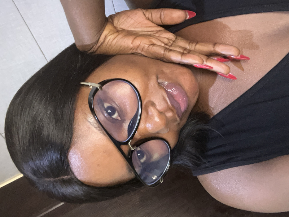 A close-up portrait features an adult female with a dark complexion, long dark hair parted at the crown, and wearing black aviator-style glasses with clear lenses. Her eyes are visible through the lenses, directed towards the viewer. Her lips are coated with a glossy, light-toned product. She is dressed in a black sleeveless garment. Her left hand is raised, with fingers splayed and resting against her right cheek/jawline. Her fingernails are long, pointed, and painted red. The background is an indistinct light-colored, possibly tiled or paneled, interior wall. This scene is located in Dzhidu, Nigeria.