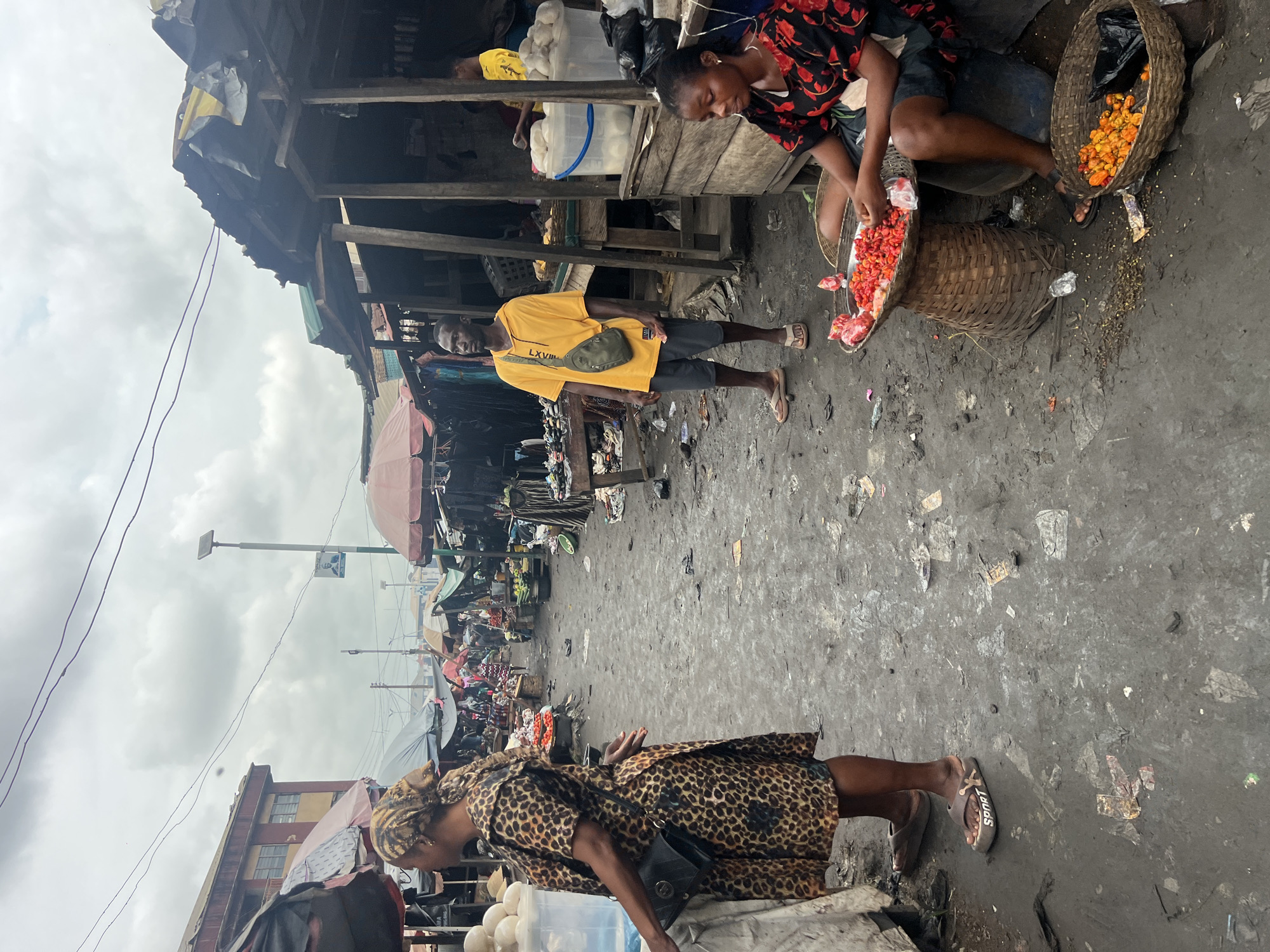 The image depicts a bustling outdoor market scene in Nkpor, Nigeria. A woman wearing a leopard-print dress and headscarf stands on the left, facing away from the camera, with a black purse slung over her shoulder. In the center, a man in a yellow t-shirt and grey shorts stands, looking to his left. To his right, a woman is seated on a low stool, tending to baskets of red and orange peppers. The market is characterized by makeshift stalls with corrugated iron roofs and wooden structures, displaying various goods, including clothing and produce. The ground is dirt, littered with debris. The sky is overcast, suggesting a cloudy day.