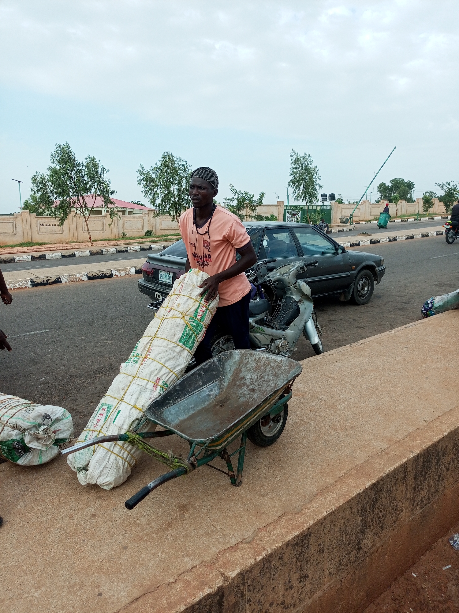 A man in a peach-colored t-shirt and a dark patterned headwrap is standing by a road, holding onto a large, white, cylindrical bundle wrapped in plastic and secured with yellow ropes. The bundle is resting on the handles of a green wheelbarrow. Another similar bundle is partially visible to the left of the wheelbarrow. Behind the man and the wheelbarrow, a dark gray sedan is parked, and a silver motorcycle is leaning against it. The scene takes place on the side of a paved road with a curb and a sidewalk. In the background, there are trees, a beige wall with ornate designs, and buildings. The sky is overcast.