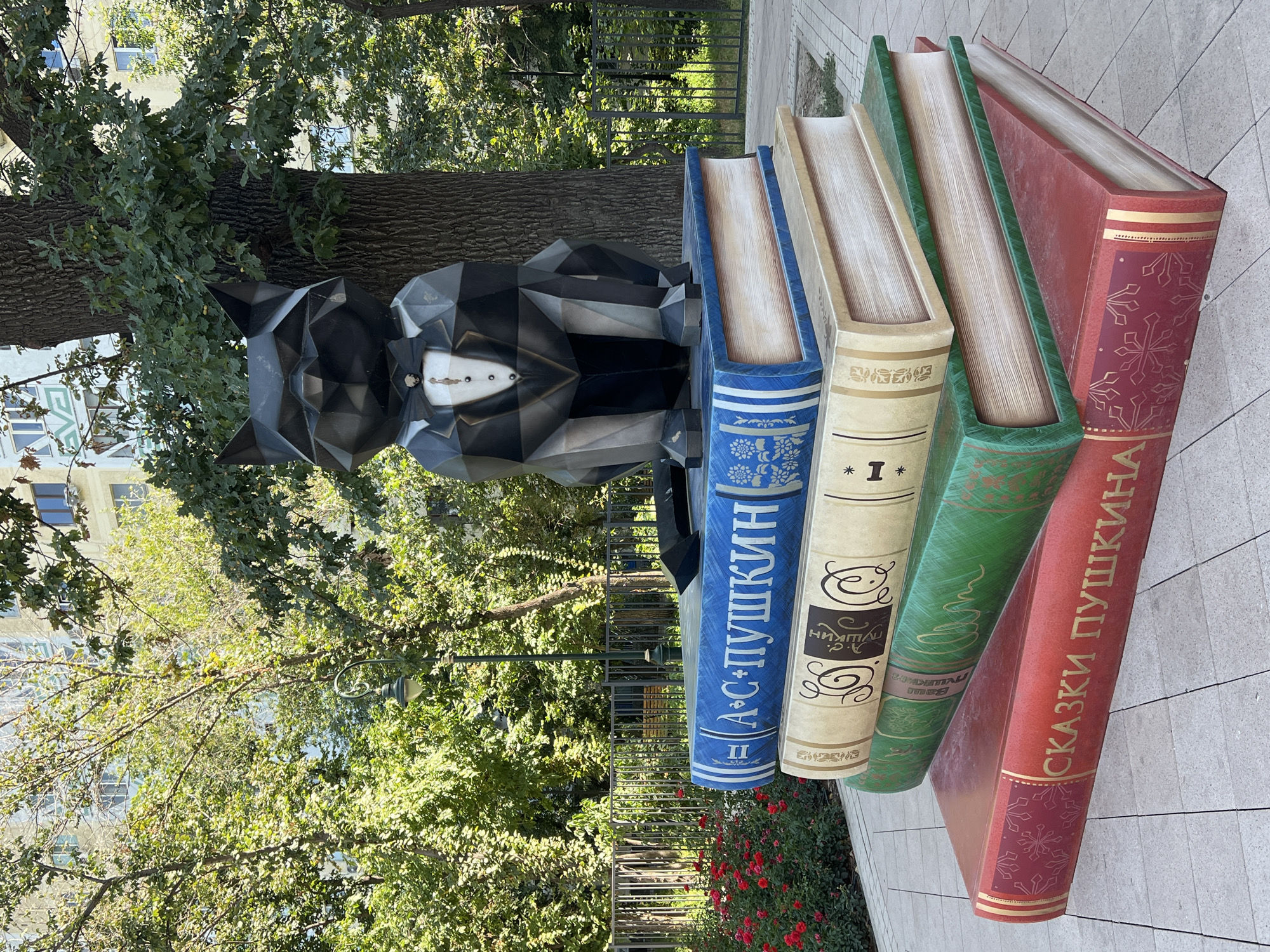 A polygonal sculpture of a black cat wearing a tuxedo sits atop a stack of oversized books in a park setting. The cat sculpture is geometrically stylized and features a white shirt with a bow tie and a brooch. The books are arranged in a pyramid shape, with the largest book at the bottom. The spines of the books are decorated with what appear to be Cyrillic text and floral or snowflake patterns. The top book is blue and has the text "А. С. ПУШКИН" (A. S. Pushkin). The book below it is tan with ornate calligraphy and the text "ПУШКИН" (Pushkin). The third book is green with similar ornate text, and the bottom book is red with gold lettering that reads "СКАЗКИ ПУШКИНА" (Pushkin's Fairy Tales).
The background consists of lush green trees, some of which are large and mature, with a building visible through the foliage. A metal fence runs horizontally behind the books, and a lamppost is visible to the left of the cat sculpture. Red flowers are blooming in a garden bed to the far left. The ground is paved with light-colored square tiles. The image appears to be taken in daylight, with sunlight casting shadows.
This sculpture is located in Almaty, Kazakhstan, specifically in the park named after Alexander Pushkin, which is located near the Almaty State Kazakh Drama Theatre.