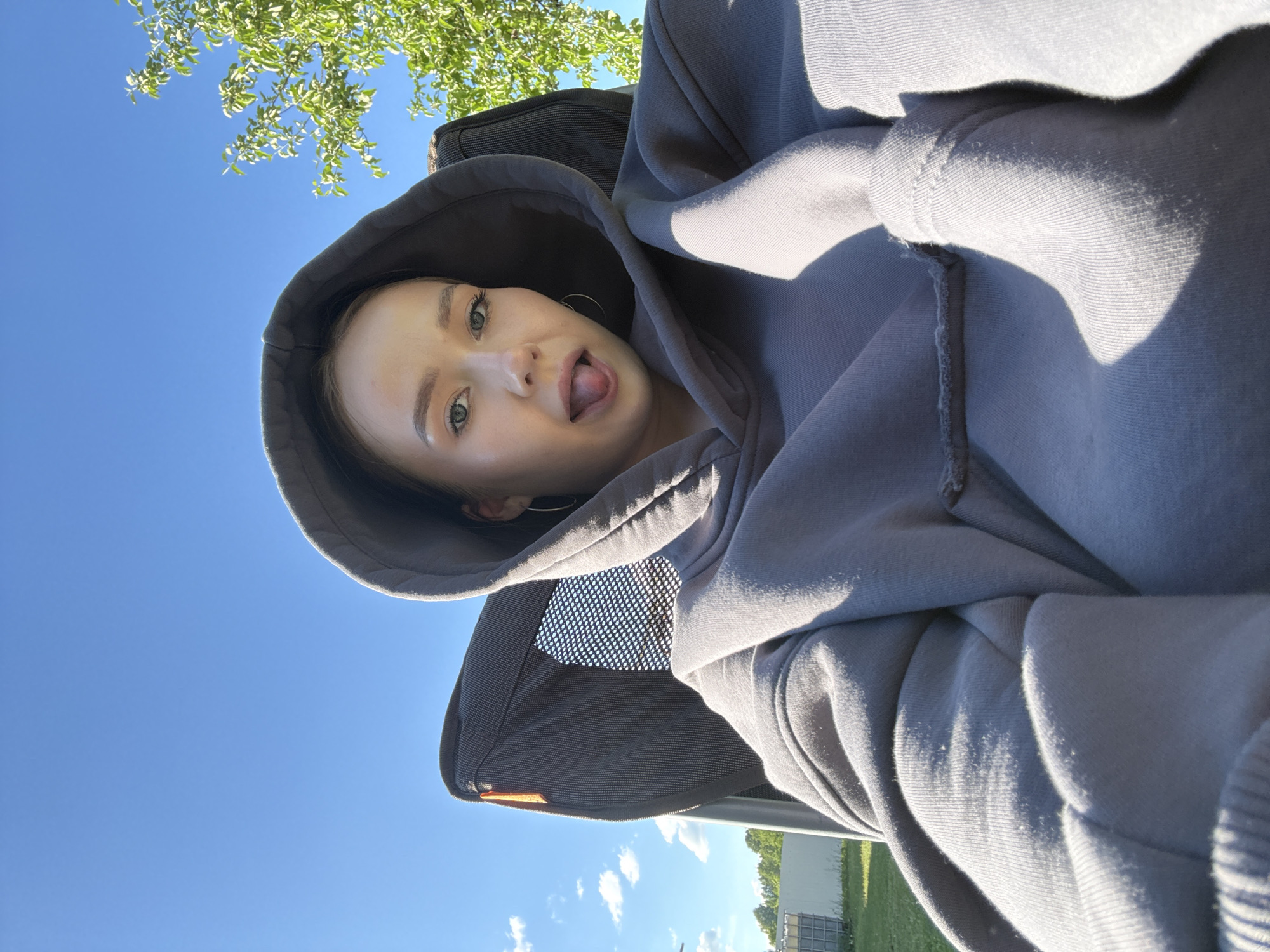 The image depicts a young woman wearing a gray hoodie, her hood up, standing outdoors in Zhigalki, Russia. She is sticking her tongue out at the camera. Above her head are the leaves of a tree, and in the background is a clear blue sky. In the bottom part of the image, some buildings are visible in the distance. The photo appears to be taken from a low angle.