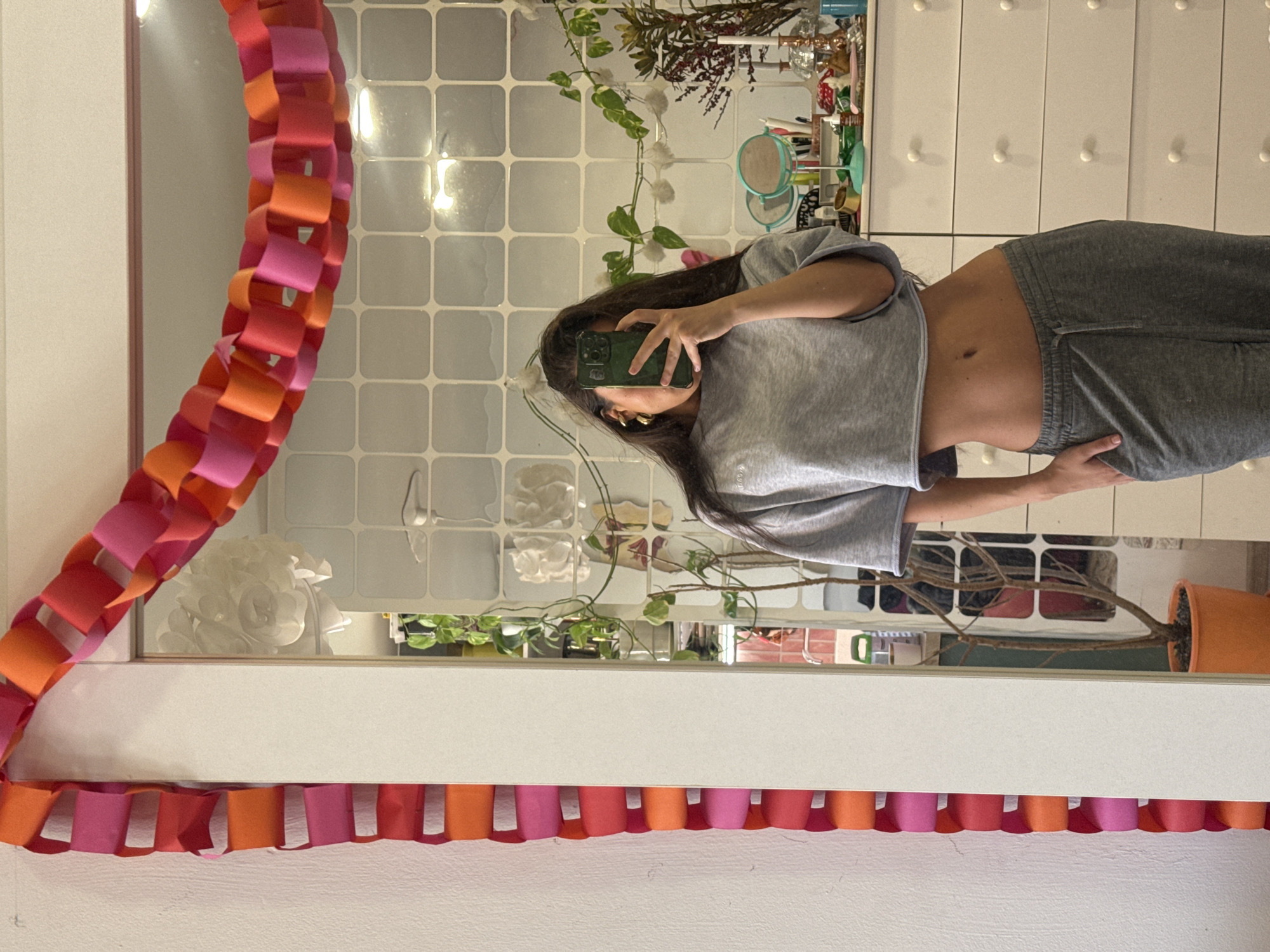A person in a grey crop top and grey sweatpants stands in front of a mirror taking a selfie with a green phone. The mirror is decorated with a paper chain of pink and orange links. The background includes a tiled wall, a white dresser, and plants. The location is not discernible.