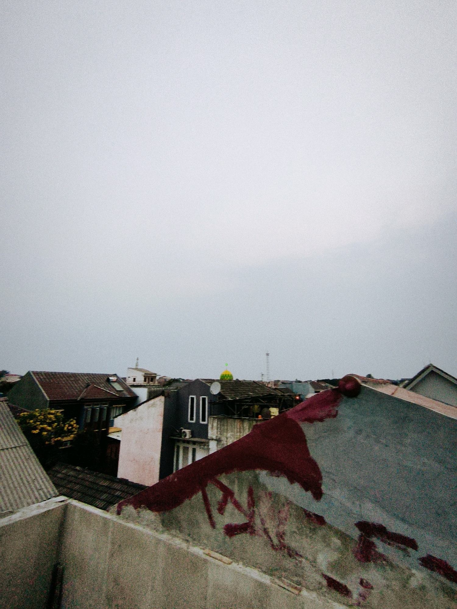 The image depicts a view of rooftops in the city of Bekasi, Indonesia. In the foreground, a textured concrete ledge is visible, leading to a sloped, weathered roof. This roof is predominantly gray with significant patches of dark red discoloration, appearing to be paint or rust stains, with some graffiti-like red markings. Beyond this, a cluster of houses and buildings fills the middle ground. Notable structures include houses with tiled roofs, one with a white facade and dark rectangular windows, and another with a dark gray exterior. A bright green dome, possibly belonging to a mosque, is visible in the distance, with a tall, slender white tower next to it. The sky above is overcast and pale gray, suggesting either early morning, late afternoon, or a cloudy day. The scene is residential, showing the typical urban landscape of Bekasi with its densely packed houses and varied building materials. There are no people or animals visible in the frame.