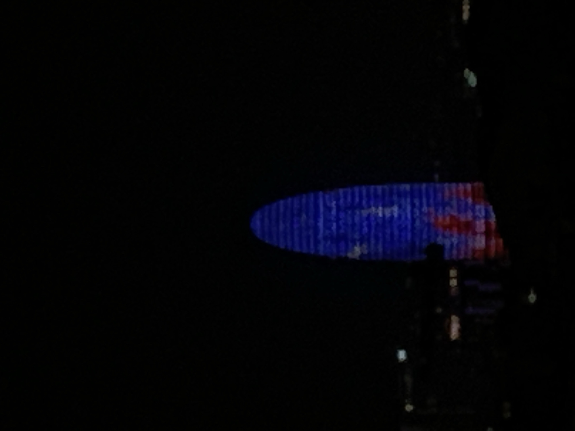 The image shows a building with a large, illuminated oval display on its side, likely located in Barcelona, Spain. The display features a pattern with a blue background and red elements, resembling an advertisement or artistic projection. Below the display, the building has multiple floors illuminated with lights, suggesting it is occupied. The surrounding environment is dark, indicating it is night, and the image appears to have been taken from a distance. There is a person walking or standing nearby the building. The exact nature of the display and the identity of the building are not discernible from the image.