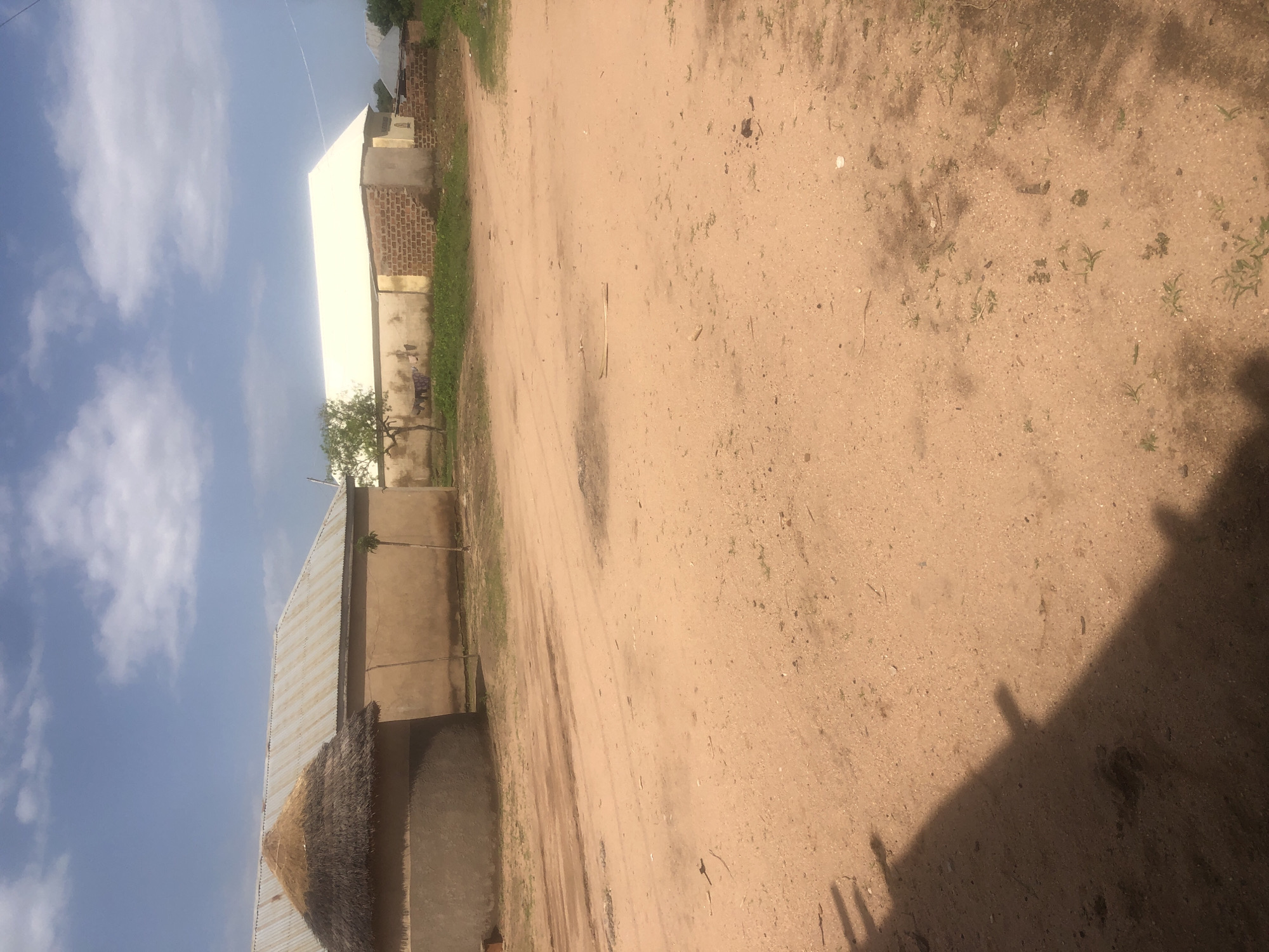 The image depicts a dirt road in a village or rural area, likely on the outskirts of Jalingo, Nigeria, under a partly cloudy sky. Two buildings are visible on the left and in the center-right of the frame. The building on the left has a thatched conical roof and a corrugated metal roof, with walls of what appears to be mud or concrete. The building on the right has a white, pitched corrugated metal roof, with lower walls constructed from brick and a lighter colored material. A small tree stands in front of this building. The road is predominantly sandy and reddish-brown, with some tire tracks and debris scattered across its surface. The shadow of an unseen object, possibly a fence or structure, stretches across the lower left portion of the image. There are no people or animals immediately visible in the scene. The overall environment suggests a warm, dry climate.