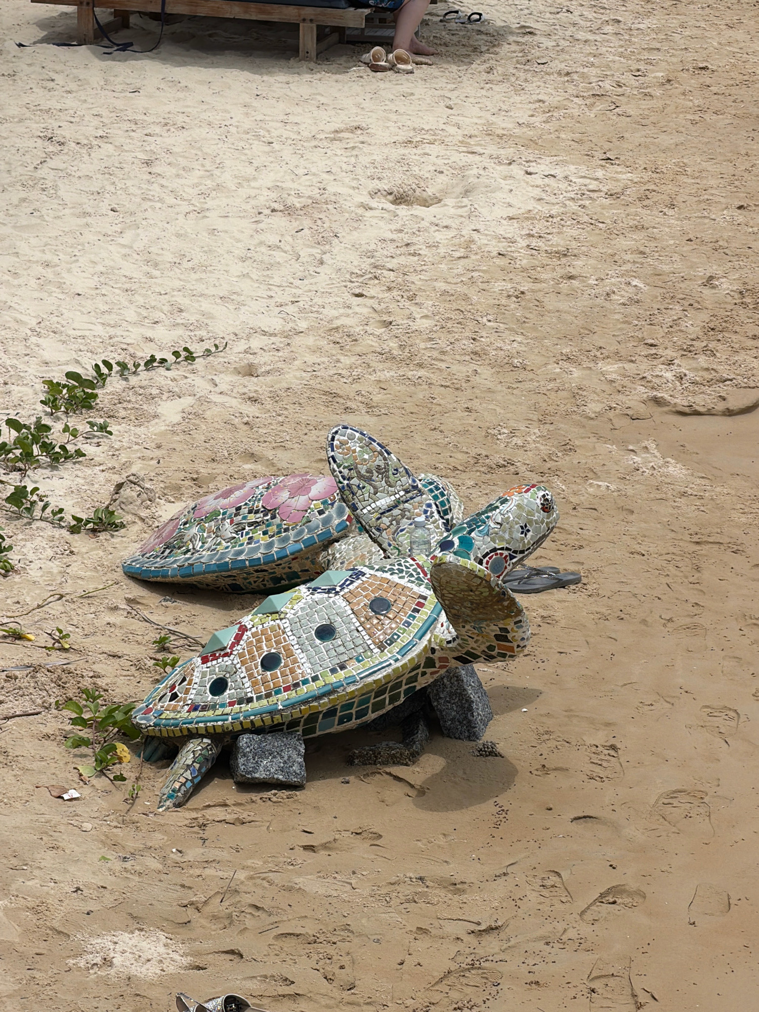 The image shows a beach in Nha Trang, Vietnam, with several mosaic turtle sculptures on the sand. The sand is light brown and there are footprints visible. To the left, there is some green vegetation growing. In the background, a wooden beach lounger is partially visible, and a person's legs and feet are resting on it, wearing sandals. The mosaic turtles are colorful and detailed, with one larger turtle in the foreground and two smaller ones behind it. One of the smaller turtles has pink floral patterns. A pair of dark flip-flops is placed near one of the turtles. The scene suggests a relaxed beach environment.