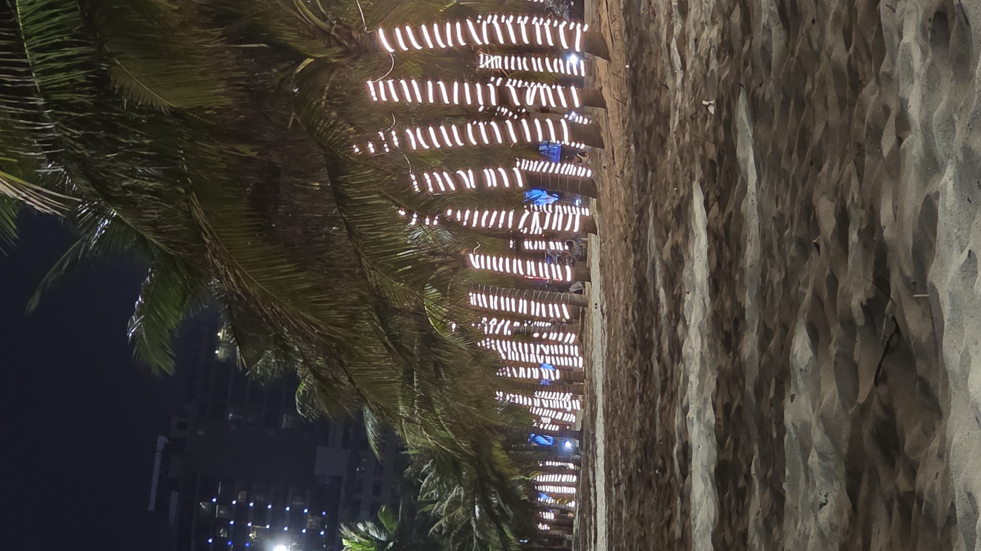 This image depicts a nighttime scene along a sandy beachfront in Nha Trang, Vietnam, consistent with the promenade often found along Tran Phu Street. The foreground consists of dark, undulating sand, showing varying textures and slight ridges. A distinct line of numerous palm trees forms the midground, with each tree trunk extensively wrapped in horizontal bands of bright white LED strip lights, creating a prominent striped illumination. Beyond these primary lit trees, additional blue ambient lighting is visible, and several indistinct human figures are discernible standing or seated between and behind the palm trees, indicating presence and activity. The distant background features tall, multi-story urban buildings with scattered illuminated windows, set against a dark night sky.
