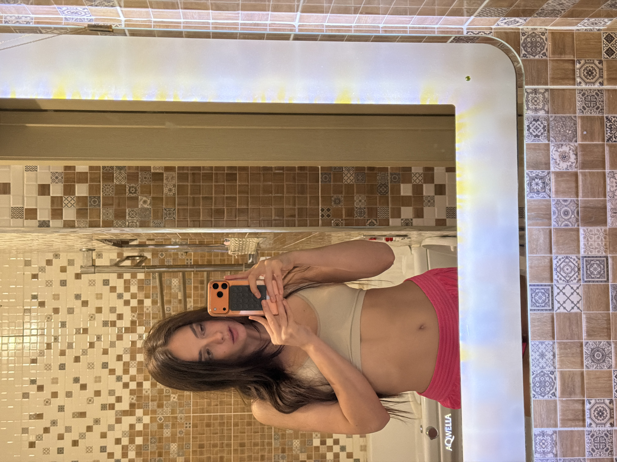 A person is taking a mirror selfie in what appears to be a bathroom. The person is wearing a beige sports bra and pink shorts. They are holding an orange smartphone to take the photo. The bathroom has tiled walls with a mosaic pattern of brown and beige squares. A large, illuminated mirror is the central focus, reflecting the person and the bathroom surroundings. The lighting from the mirror is bright and casts a glow. The image is vertically oriented.