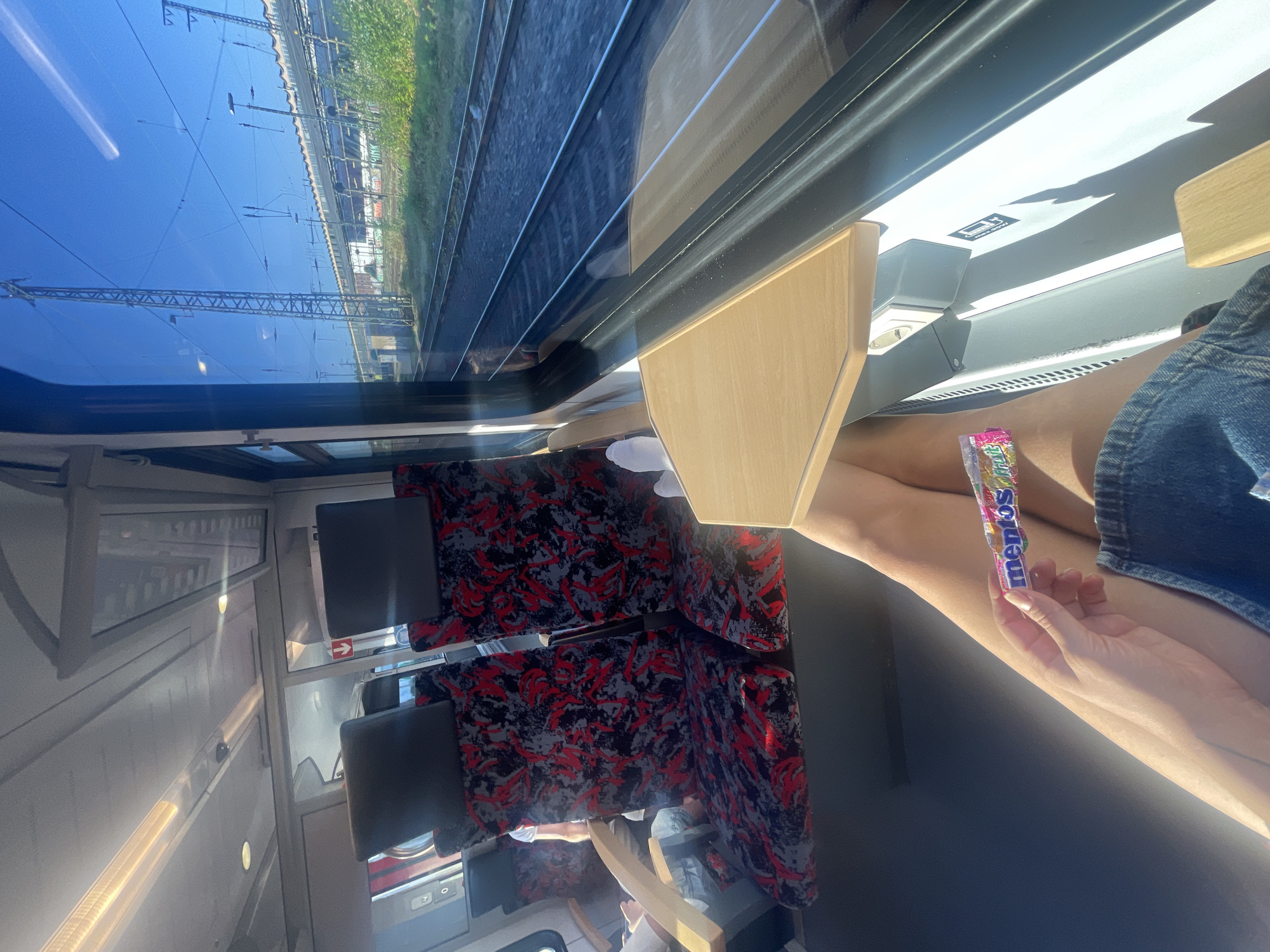 The image depicts the interior of a passenger train, with a focus on a person's legs and hand holding a pack of Mentos candies. The person is seated on a red and grey patterned seat, wearing denim shorts. A small wooden tray table is extended in front of them. The train car's interior is visible, including other seats and the ceiling. Through the window, train tracks and overhead power lines are visible against a clear blue sky, suggesting an outdoor environment near railway infrastructure. The image does not contain any information that specifically places it in the city of Hagen, Germany.