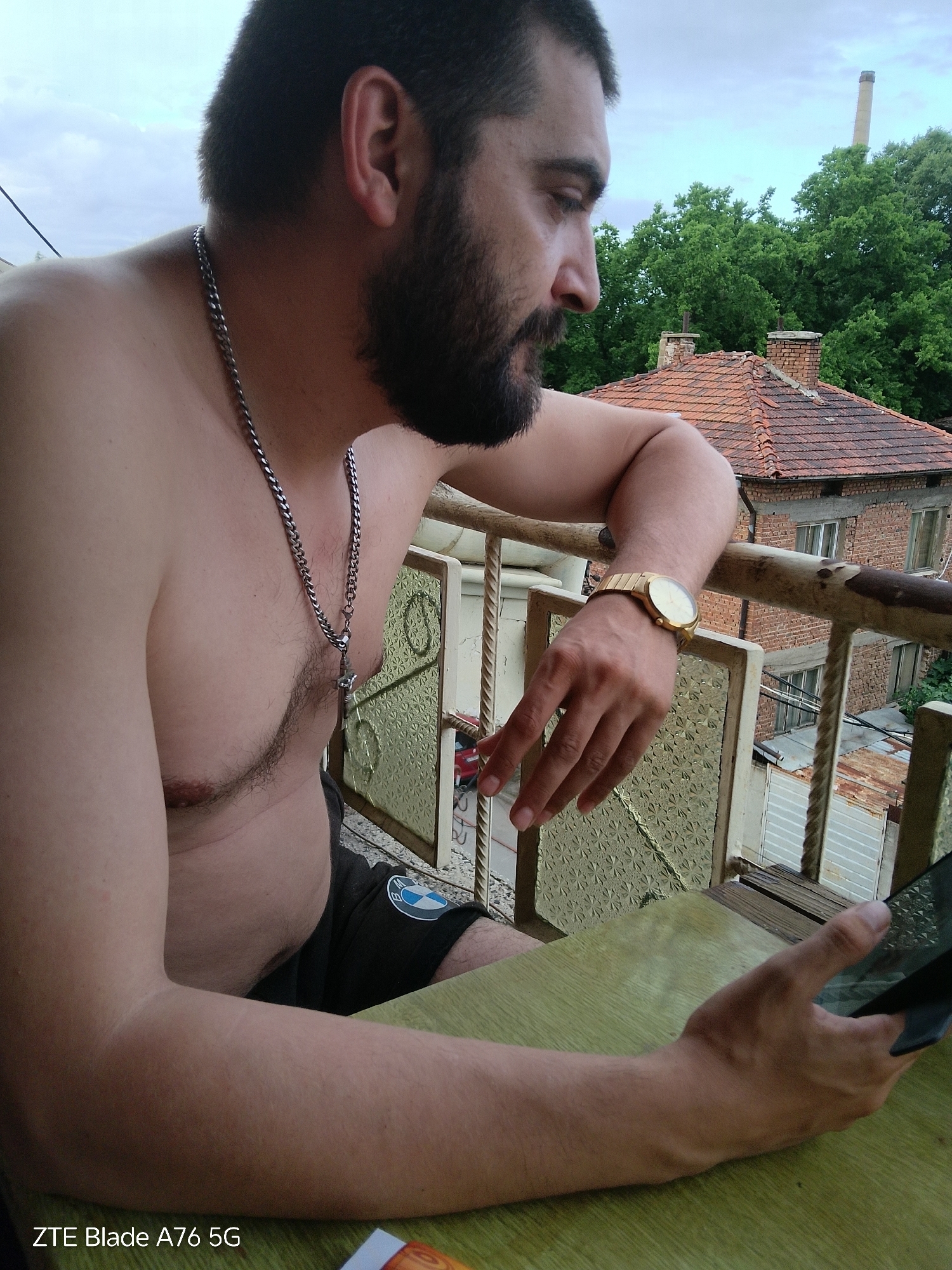 A man with a beard is sitting on a balcony, looking at his phone. He is shirtless and wearing a silver chain necklace and a gold watch. In the background, there are brick houses and trees. The photo was taken in Stara Zagora, Bulgaria.