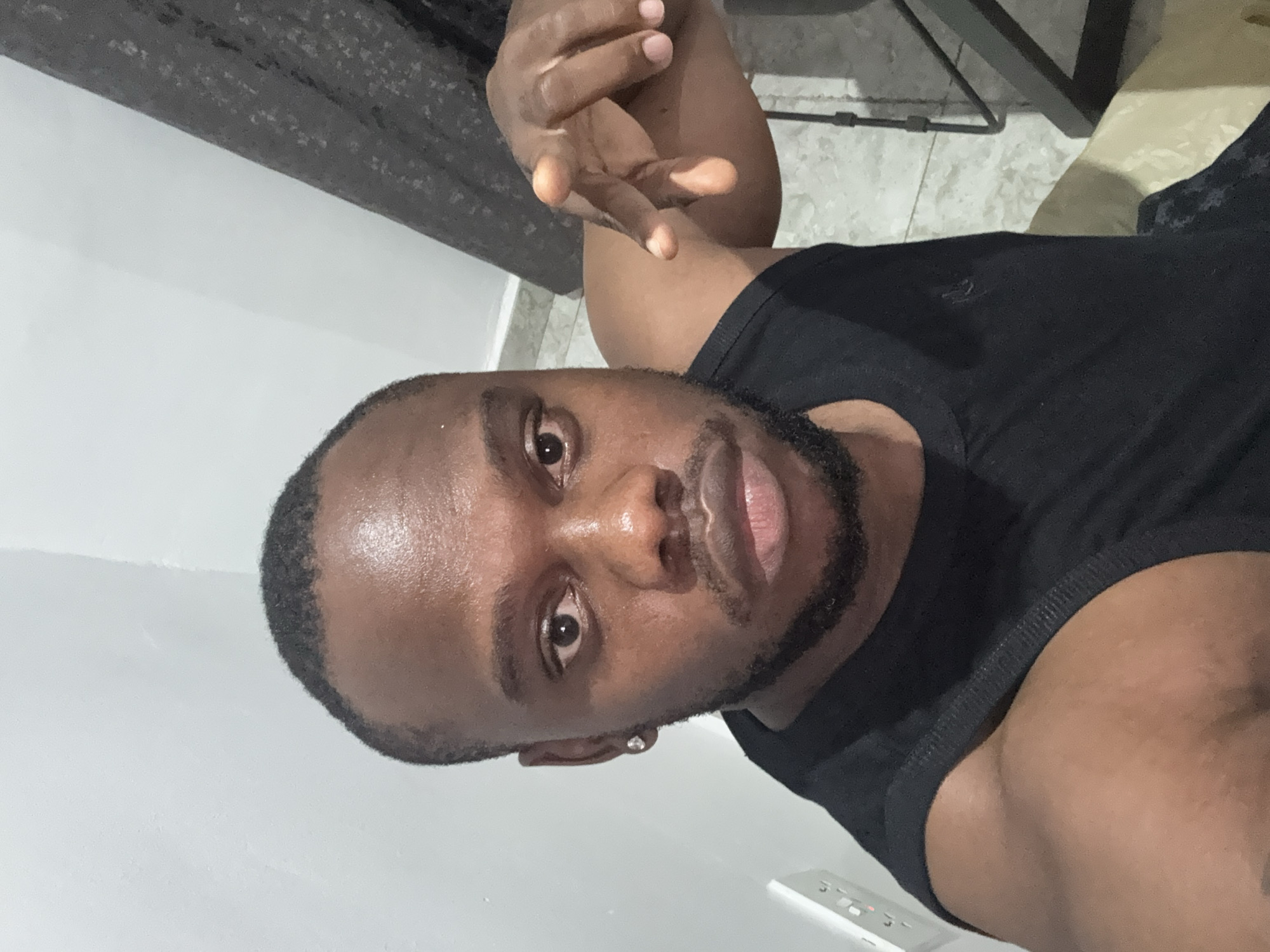 An indoor selfie captures a dark-skinned man with short dark hair and a neatly trimmed beard, located in Iddo Tudunwada, Nigeria. He is wearing a black crew-neck t-shirt and has a small, round, silver-toned stud earring in his left ear. The man is looking directly at the camera with a neutral expression, his lips slightly parted. His right hand is raised and partially open, with fingers spread in a relaxed gesture.
The background primarily features a plain, light-colored wall. To the top right, a dark, textured surface, possibly furniture or a patterned wall, is visible. The lower right portion of the image shows a light-colored tiled floor with dark furniture legs or a stand. A white rectangular light switch, with what appears to be two buttons or switches, is visible on the wall in the bottom right corner. The lighting suggests an indoor setting, with reflections indicating some light source on the man's forehead and nose. No visible text is present.