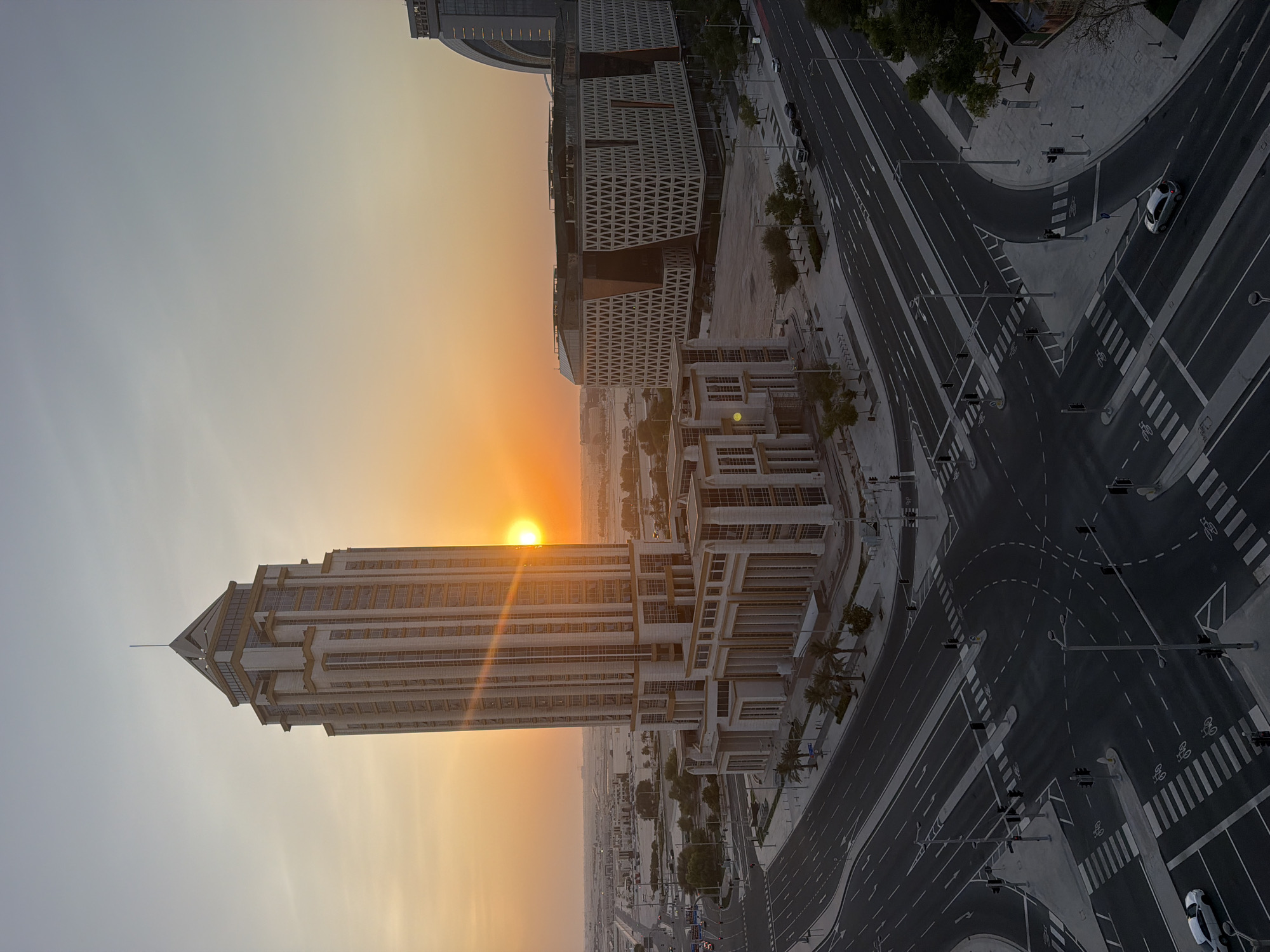 The image captures a broad aerial view of a modern urban intersection in Lusail, Qatar, at sunset. The foreground is dominated by the dark asphalt of a complex intersection with multiple lanes, pedestrian crossings, and traffic signals. Several cars, including a white sedan, are visible on the roads. Palm trees and streetlights line the sidewalks.
Dominating the mid-ground is a tall, multi-story building with a stepped architectural design, featuring numerous windows and ornate detailing. Adjacent to it is a distinctive, modern building with a facade characterized by a geometric, triangular pattern in shades of brown and white. Further in the distance, another tall, curved building is partially visible on the right.
The sunset casts a warm, orange glow across the sky, with the sun partially obscured by the taller building on the left. The horizon shows a flat, developed landscape with more buildings and infrastructure, characteristic of a rapidly developing city. The overall scene suggests a quiet evening in a well-planned, contemporary urban environment.