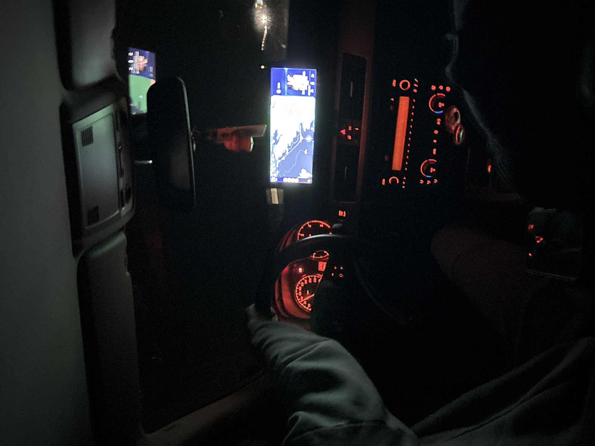 The image shows the interior of a car at night. The dashboard is visible, with a lit-up steering wheel, speedometer, and other gauges. A phone mounted on the dashboard displays a map. There is a person behind the wheel, though only part of their arm and torso are visible. The passenger side and center console area can be seen. The location cannot be confirmed with the image.