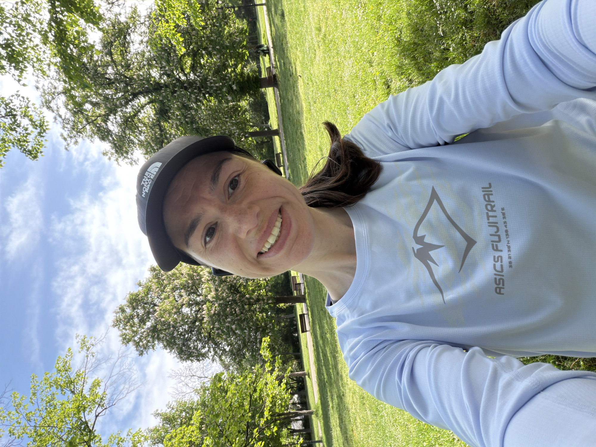 A woman is taking a selfie in a park. She is wearing a black cap with a logo, a light blue long-sleeved shirt with the words "ASICS FUJITRAIL 35 21'36"N 138 43'36"E", and has brown hair. The background includes green grass, trees with green leaves, and a blue sky with clouds. There are benches in the background. Considering the shirt's coordinates, it is unlikely to be Sofia, Bulgaria; the coordinates point to Mount Fuji in Japan. Therefore, the location cannot be confirmed as Sofia, Bulgaria based on the provided information.