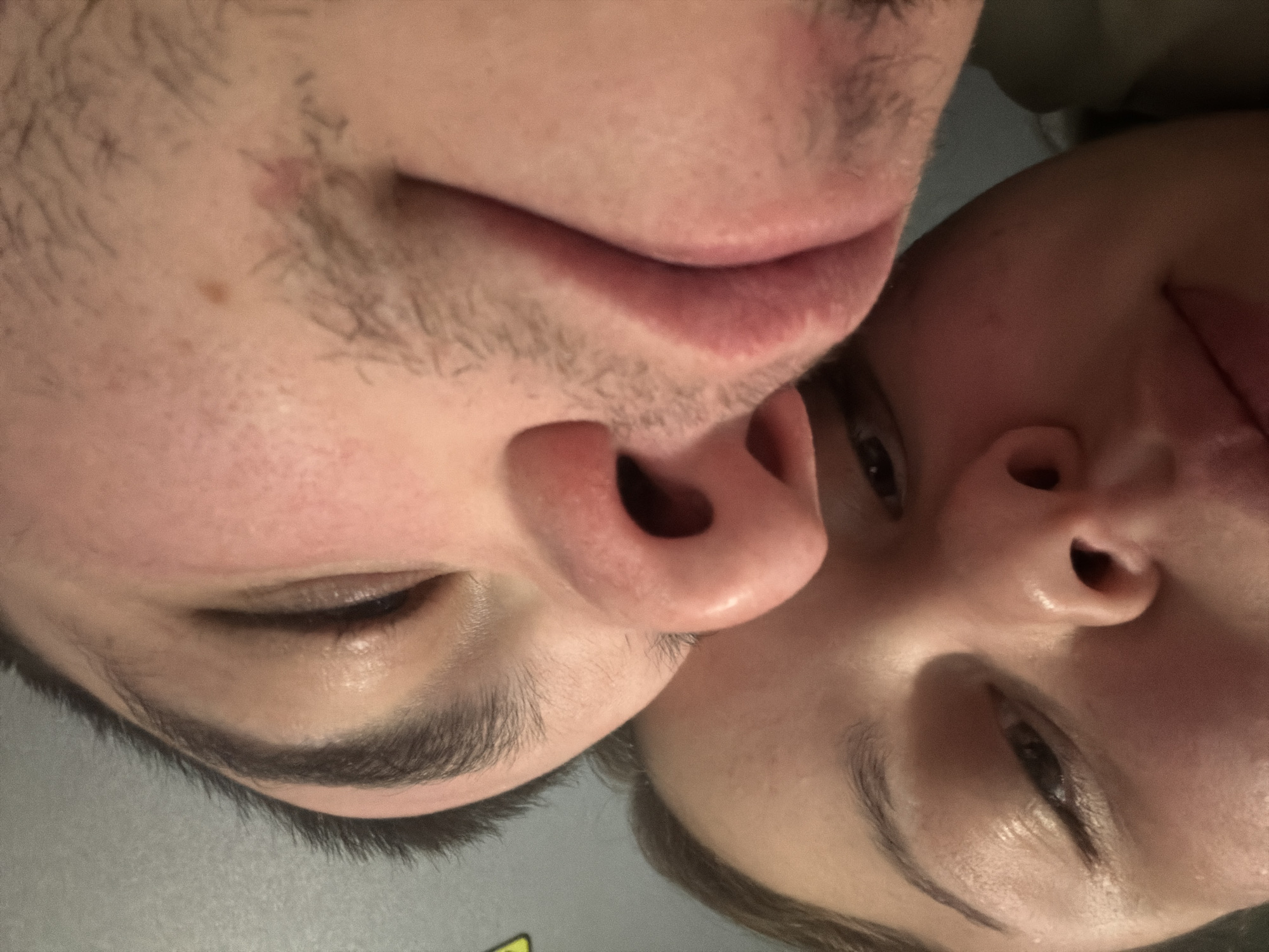 The image shows two faces closely pressed together, oriented upside down. The face on the left is male, with visible facial hair on the upper lip and chin, and a mole on the cheek. The face on the right appears to be female. Both faces have closed eyes. The background is unclear. The location, Varna, Bulgaria, is unknown from the image.