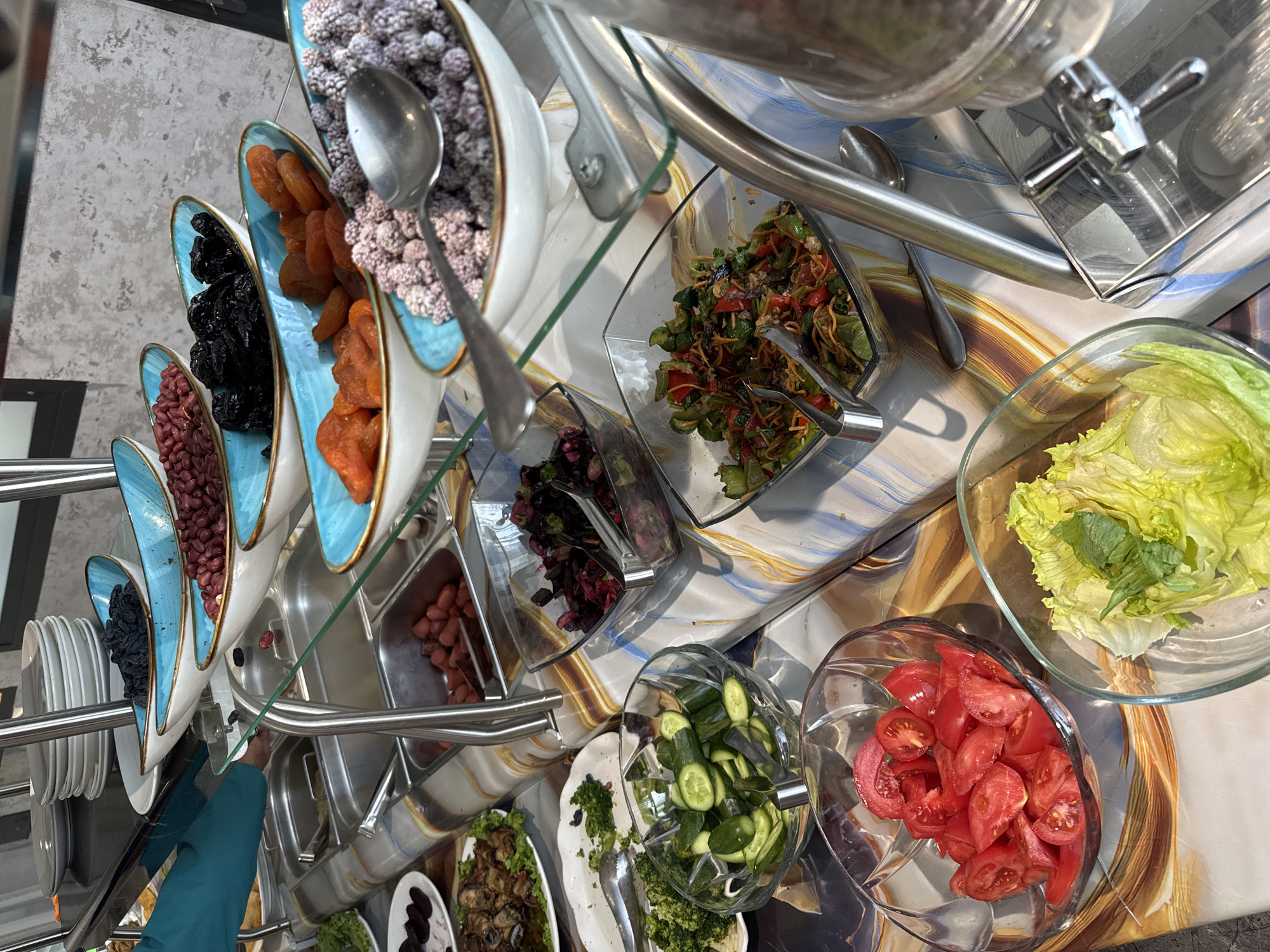 The image presents a buffet-style food arrangement, likely within a dining facility. A variety of food items are displayed in bowls and serving dishes including dried fruits, various salads, sliced cucumbers and tomatoes, and leafy greens. Serving utensils such as spoons and tongs are positioned beside each dish. The arrangement suggests a self-service setting. The location appears to be indoors. The presence of a beverage dispenser in the upper right corner indicates a potential breakfast, lunch, or dinner buffet. The exact location within Tashkent, Uzbekistan is not ascertainable from the image. There are no discernible people in the image.