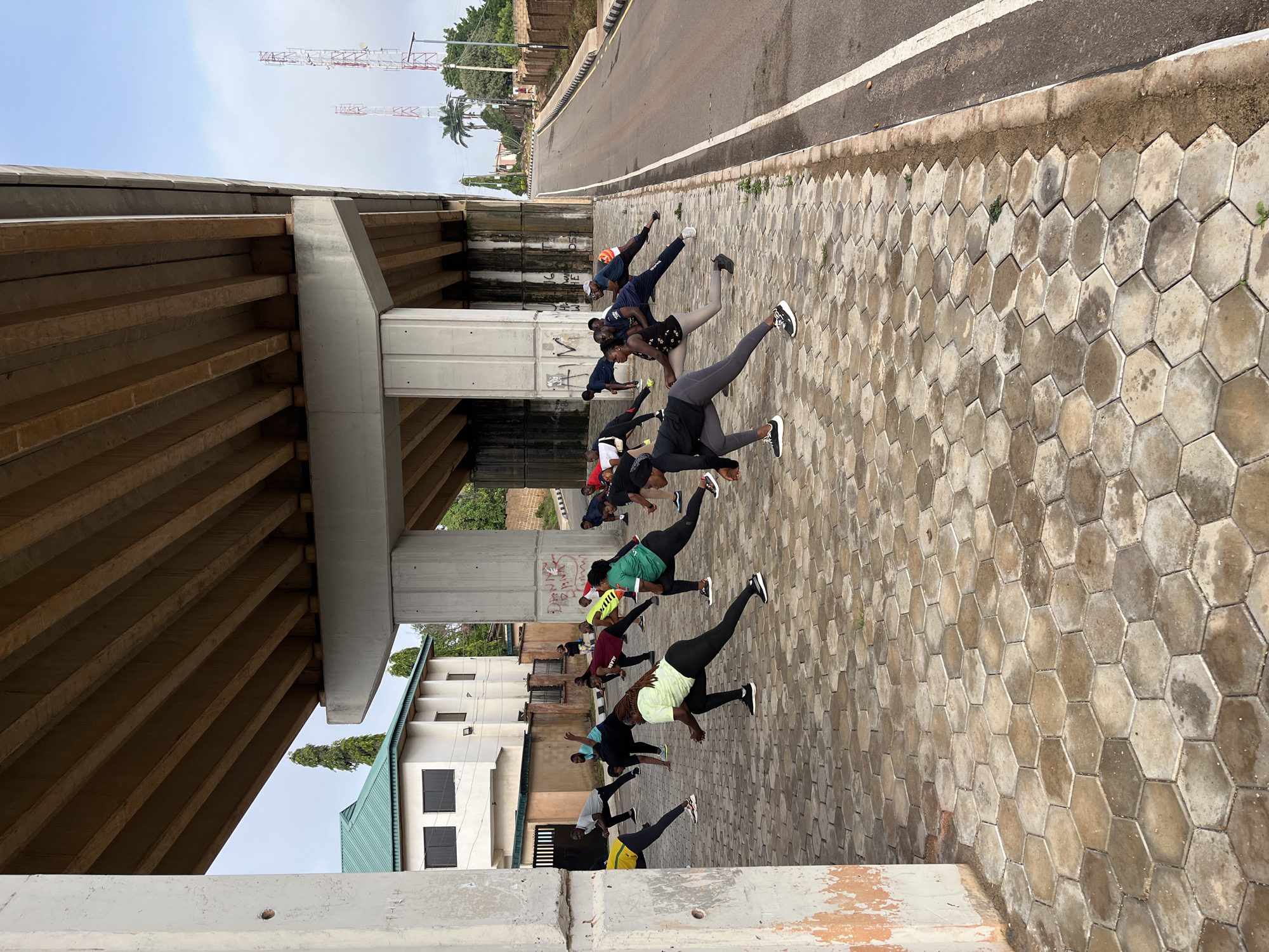 The image captures an outdoor exercise session taking place under a large, concrete overpass, likely within the city of Jos, Nigeria. A group of approximately 15 individuals, dressed in athletic wear, are engaged in various stretching exercises. The ground beneath them is paved with hexagonal concrete tiles. In the background, a white building with a green roof and dark windows is visible, along with trees and a road. Two prominent communication towers with red and white markings stand against a cloudy sky, further indicating an urban environment. The overpass supports are also made of concrete, and graffiti is present on one of the support pillars. The overall scene depicts a public space being utilized for group fitness activities.