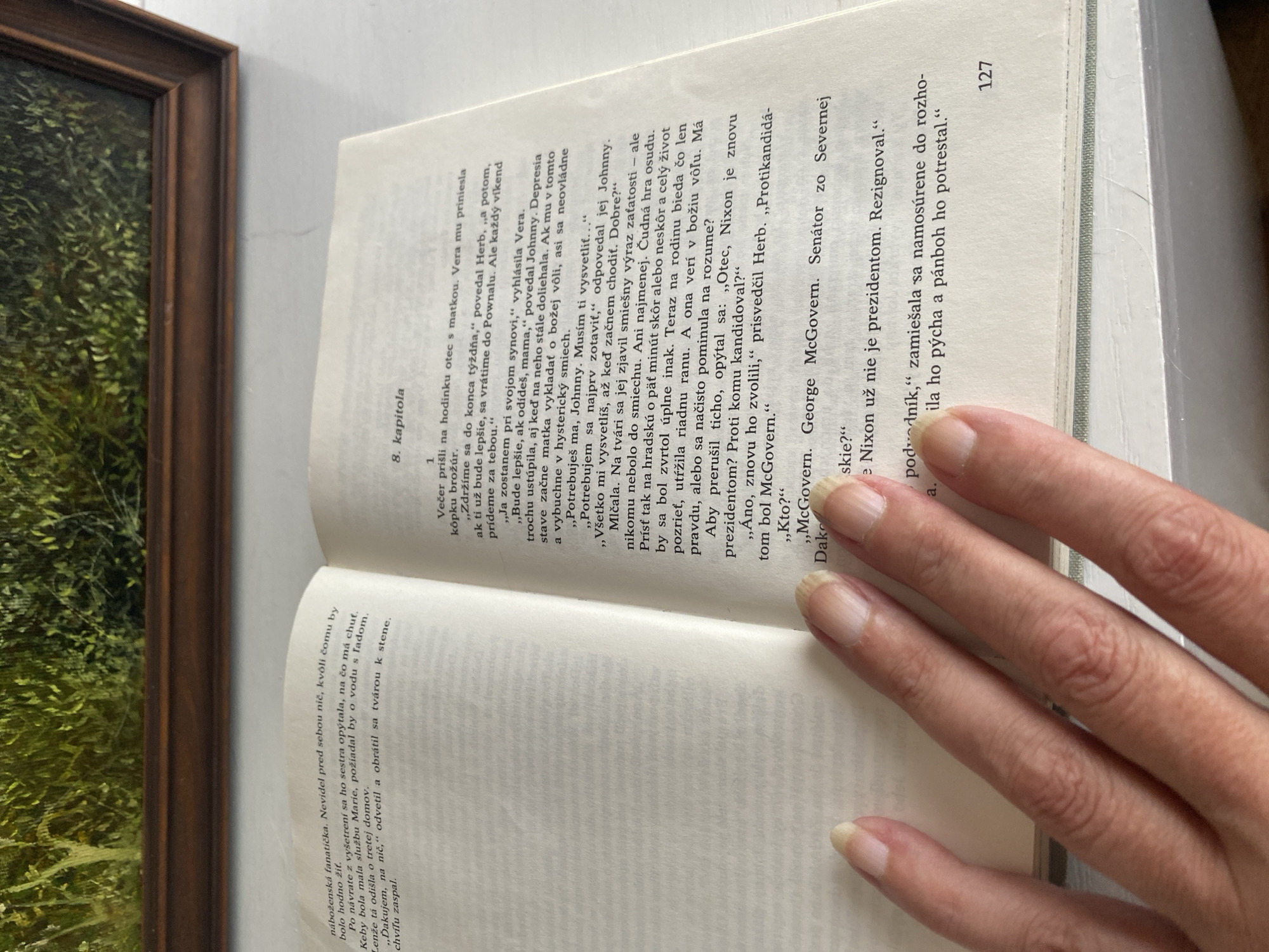 The image shows an open book with Slovak text, placed on a light-colored surface. A person's left hand, with manicured fingernails, is visible holding down the pages. The book's pages are creased and appear well-worn. The text on the right-hand page is mostly legible, with the chapter title "8. kapitola" visible. The bottom right corner of the page shows the number "127". The background suggests an indoor environment, possibly a room in a house, with a framed picture of greenery visible at the top of the image. No specific location in Bílina, Czechia is indicated.