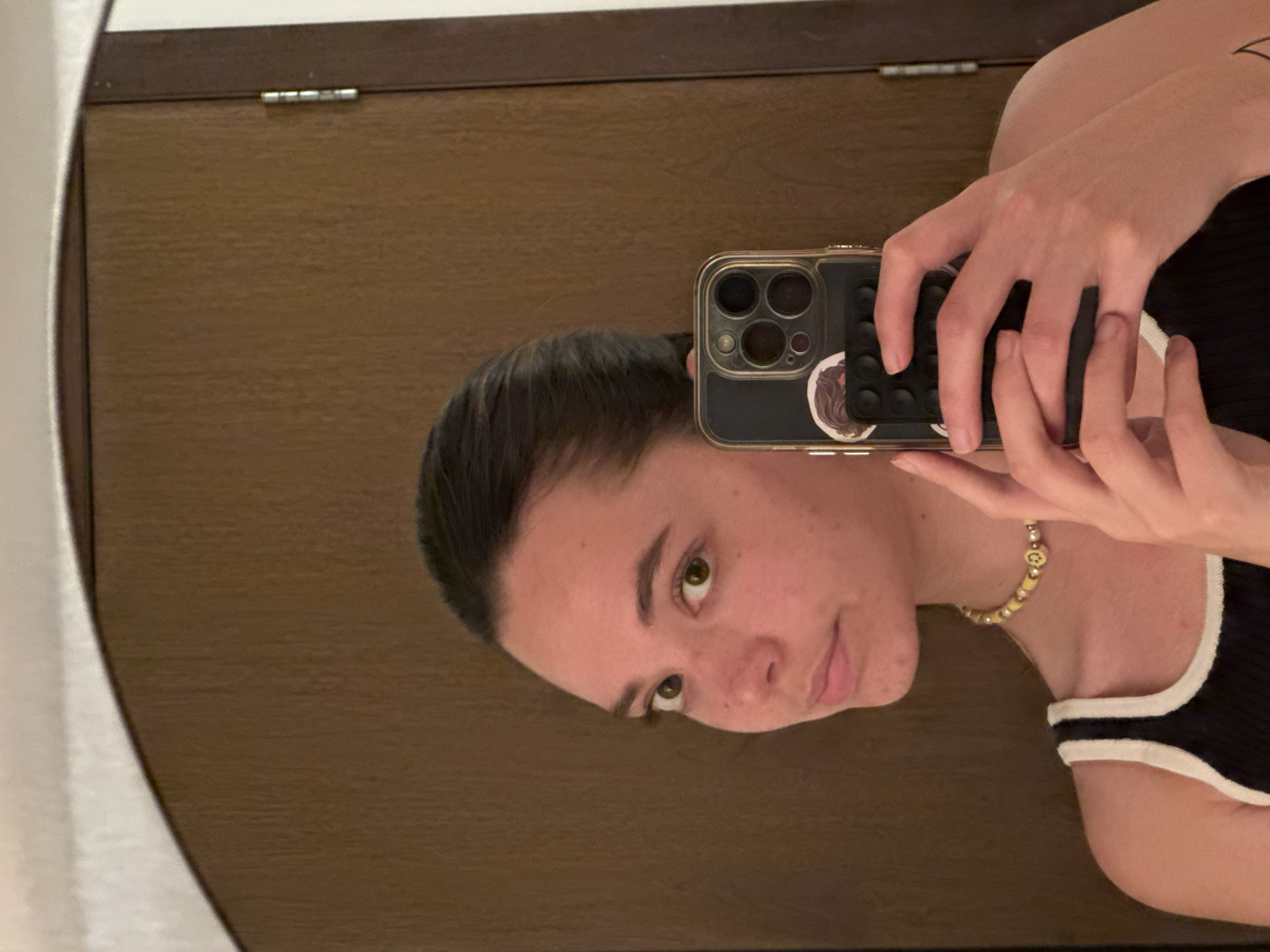 A person is taking a selfie in a mirror. The person is holding a smartphone with a dark case. The phone has multiple camera lenses visible. The person is wearing a black and white top. The background is a wooden surface with silver hardware. A gold beaded necklace is around the person's neck. The location is not discernible from the image.
