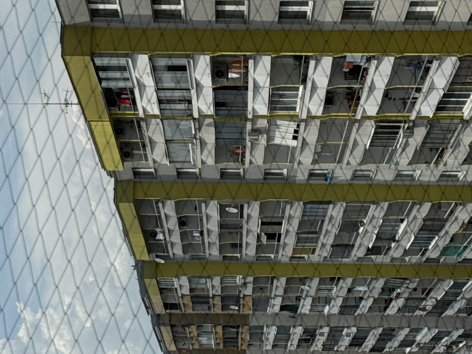 The image shows a view of a residential apartment building in Bratislava, Slovakia, seen through a diamond-patterned net. The building is tall, with multiple stories and numerous balconies. The balconies are enclosed with glass or are open, and some have laundry drying. The building's exterior is gray concrete, with bright yellow vertical accents on the corners of sections of the building. The sky is visible above the building, with scattered white clouds. The net in the foreground creates a textured, partially obscured view of the scene. No people are clearly visible.