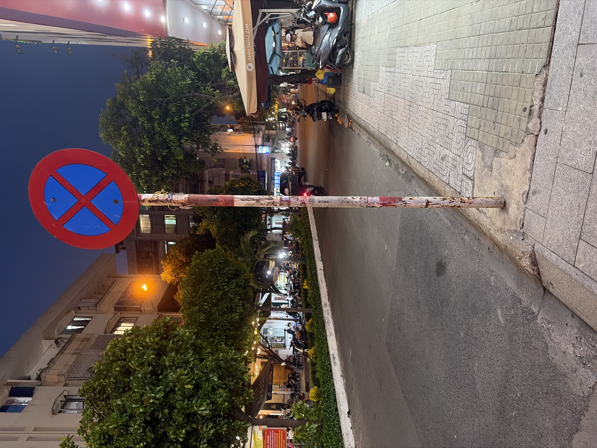 This image depicts a lively street scene at dusk or night in a city, likely in Vietnam given the architecture, vehicles, and visible text. The sky is a deep blue, with artificial lights illuminating the area.
In the foreground, a prominent red and blue "no stopping or standing" sign is visible, mounted on a weathered red and white striped metal pole. To the right of the sign and pole, an asphalt road stretches into the distance, bordered by a tiled sidewalk on the right. The sidewalk features various patterned tiles, some with geometric and swirling designs.
The street is active with traffic, mainly motorcycles, several of which are parked along the sidewalk or seen in motion on the road. Buildings line the street, many with ground-floor businesses that appear to be cafes or restaurants, indicated by awnings, outdoor seating, and decorative string lights. Lush green trees and shrubs are interspersed along the sidewalk and in front of the establishments.
A notable detail is the text "ANH NGỮ ZIM" visible on a light-colored awning, which translates to "ZIM English," suggesting an English language center or an associated cafe. The overall atmosphere is that of a busy urban environment after sunset, with a functional "no stopping" sign contrasting with the bustling social and commercial activity.