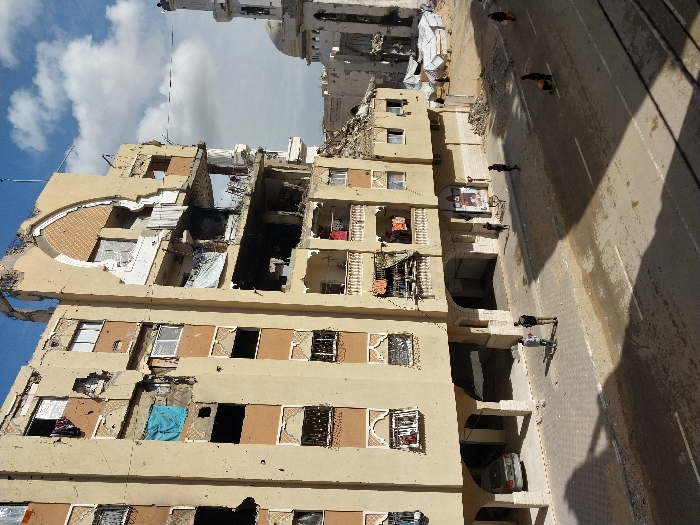 The image displays a street scene in Khan Yunis, Palestinian Territory, under a partially cloudy sky. A multi-story, light-colored building, exhibiting extensive structural damage, occupies the left and central foreground. Large sections of its facade, balconies, and internal structures are missing, revealing exposed rebar and interior spaces. Clothes hang from some remaining balconies and windows. Below the damaged building, a silver sedan is parked under its arched ground-floor section. In the background to the right, a mosque with a damaged white minaret and dome is visible amidst piles of rubble and temporary tarp shelters. On the paved street and sidewalk, approximately seven to eight individuals are present. Two men stand conversing on the sidewalk in the immediate foreground. Further down the street to the right, one person is observed pushing a cart. The street and sidewalk surfaces are marked with debris, dirt, and wet areas, with prominent shadows cast from the damaged building.